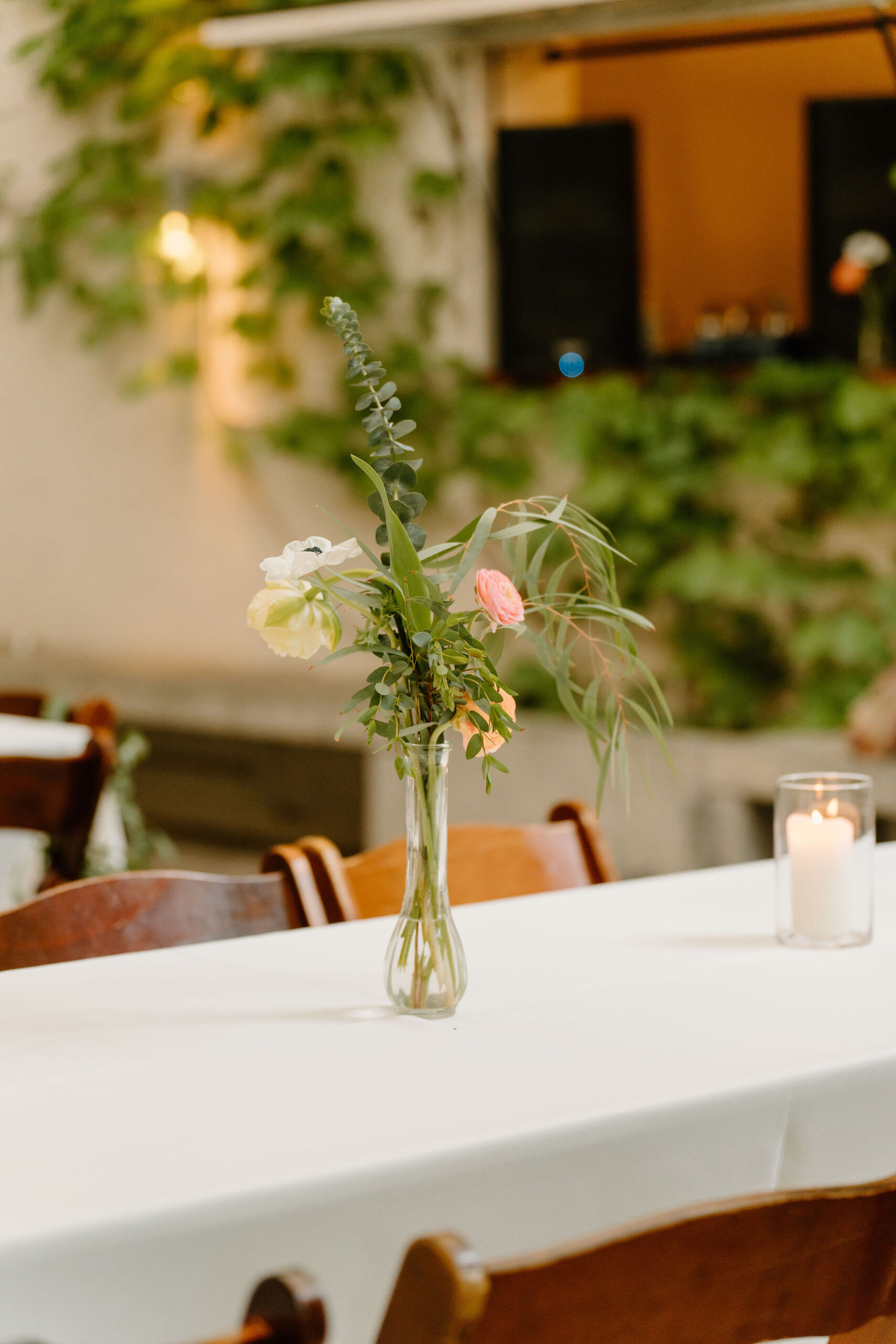 floral centerpieces in a thin tall vase, on a white tablecloth in a room filled with ivy