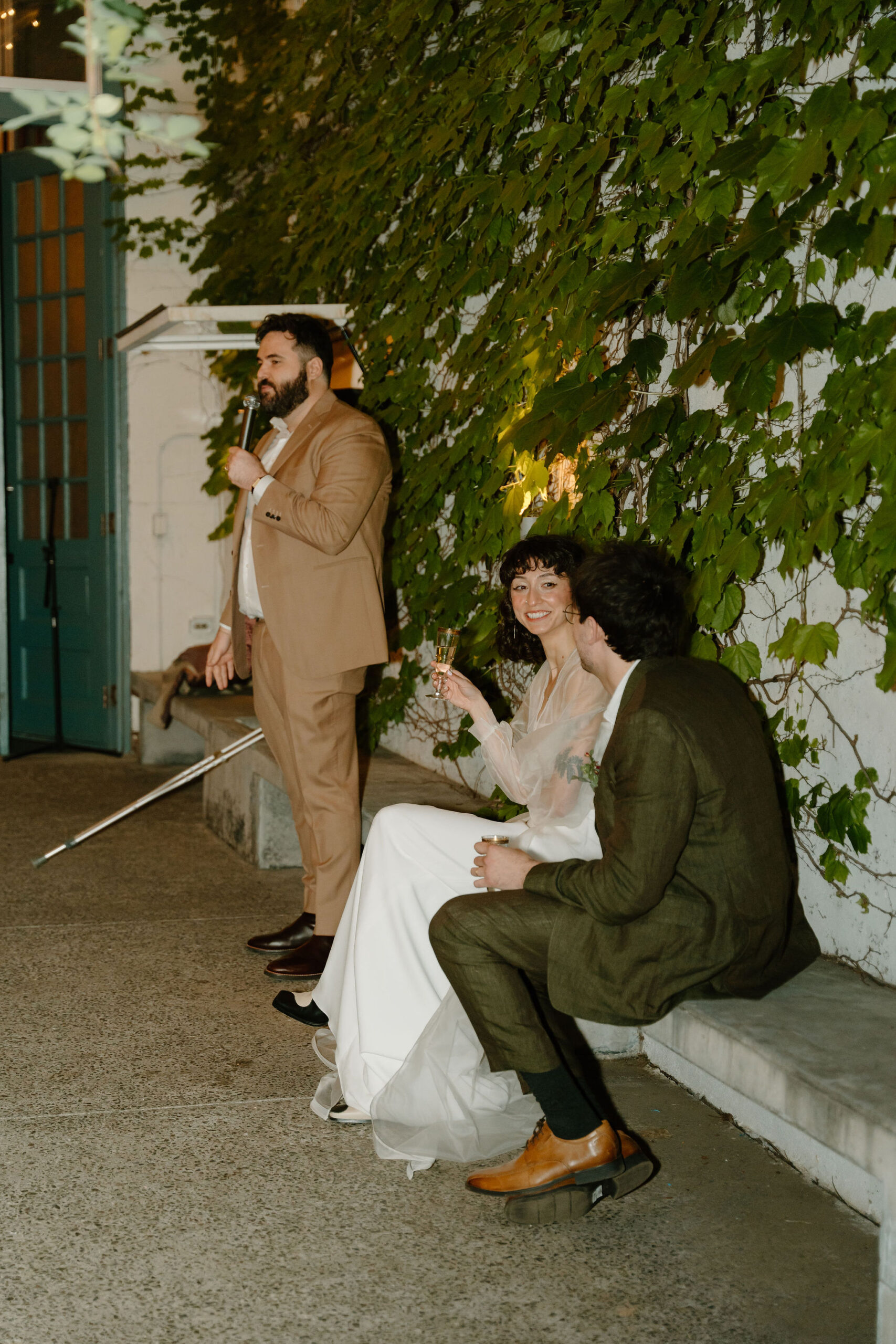 groom's friend giving a toast in front of an ivy wall, while bride and groom sit on a bench and watch