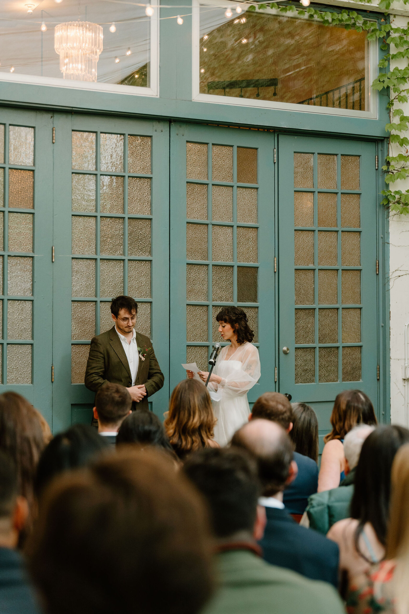 bride and groom exchanging vows during their ceremony, in front of large teal doors while their guests watch