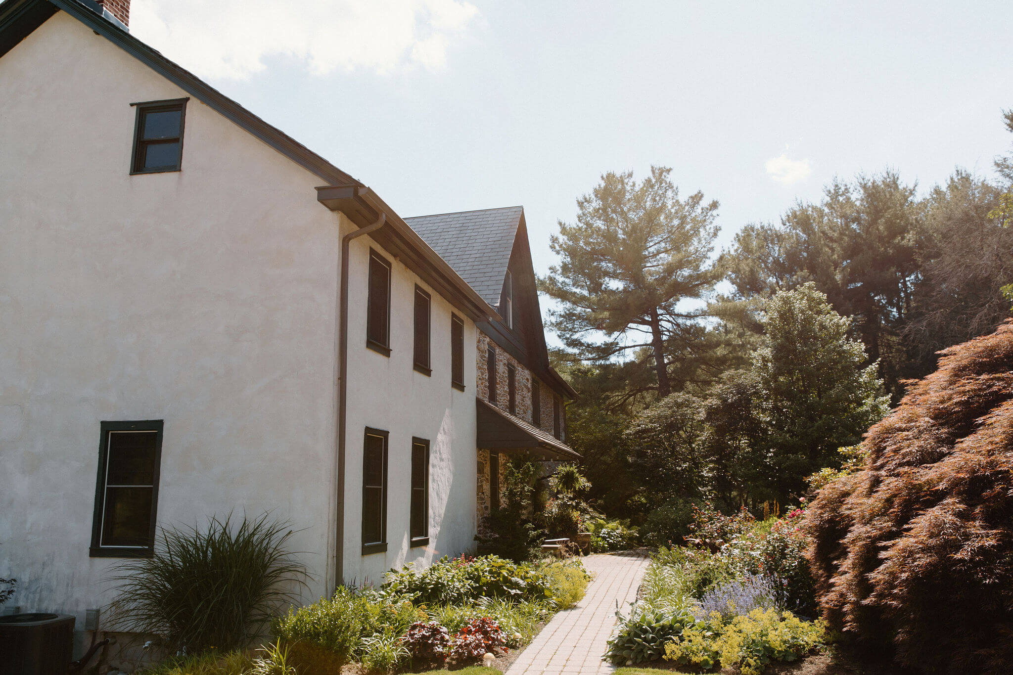 a brick path cutting through a front garden full of lush greenery and flowering bushes, leading up to the front door of a stone farm house with a stucco addition