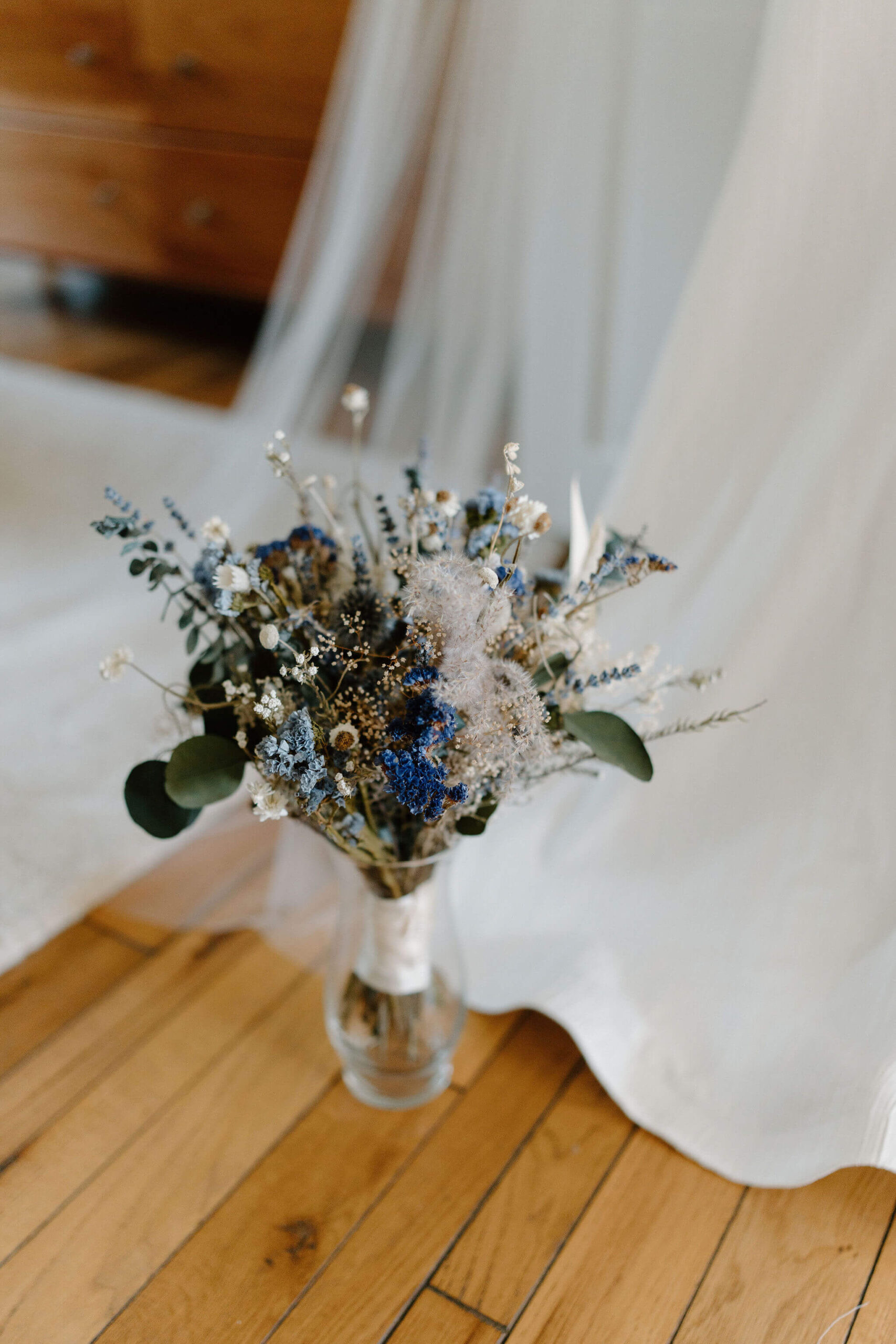 dried flower bouquet of eucalyptus, thistle, and other blue and white flowers, in a dry vase on a wooden floor, a wedding dress in the background