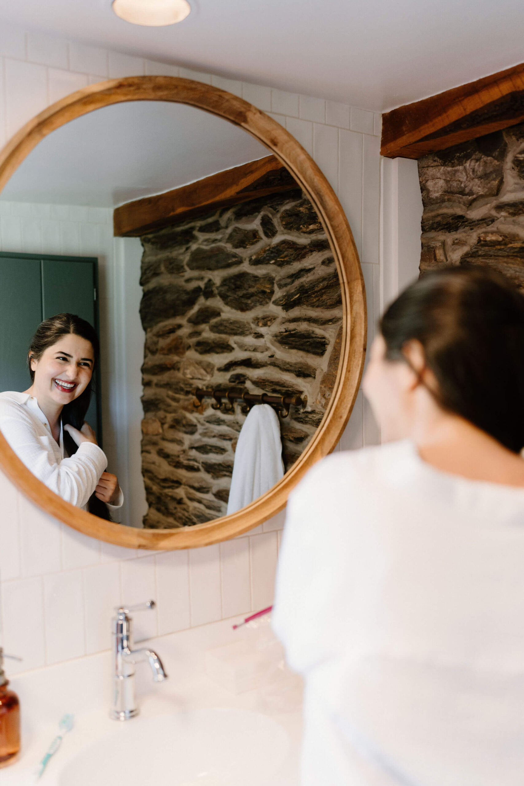 bride smiling at herself in the bathroom mirror, while styling her long brown hair