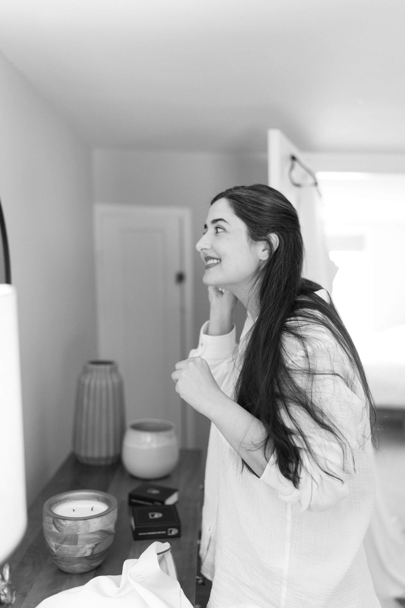 black and white image of a woman (bride, long brown hair) in a white button down shirt, looking in a bedroom mirror to put her earrings in