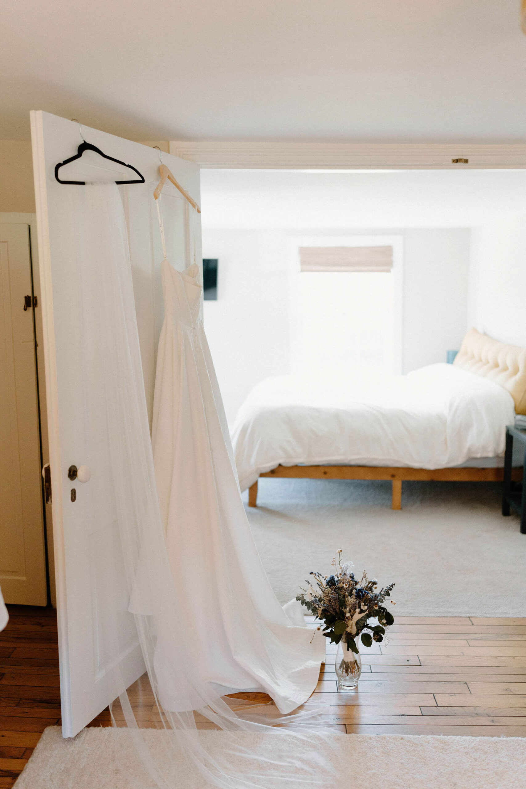 wedding dress and veil on hangers, hanging on a bedroom door. there is a vase of dried florals set on the floor next to the dress, and a made bed in the background.