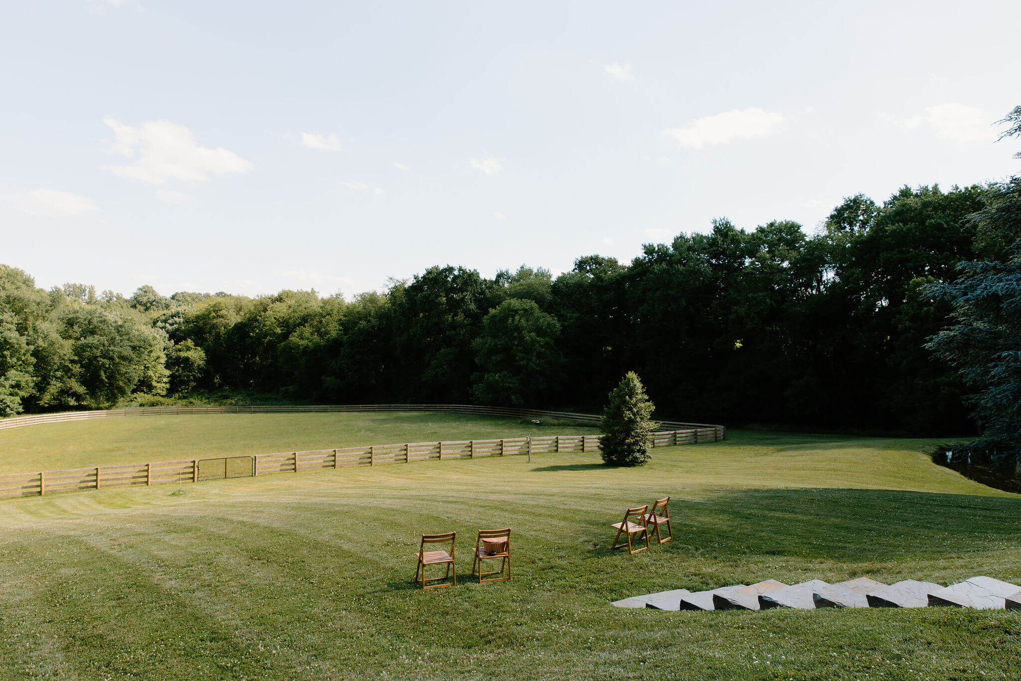 four wooden chairs placed in an open field, with a rolling hill and forest in the background. A large fence cuts through the middle of the field