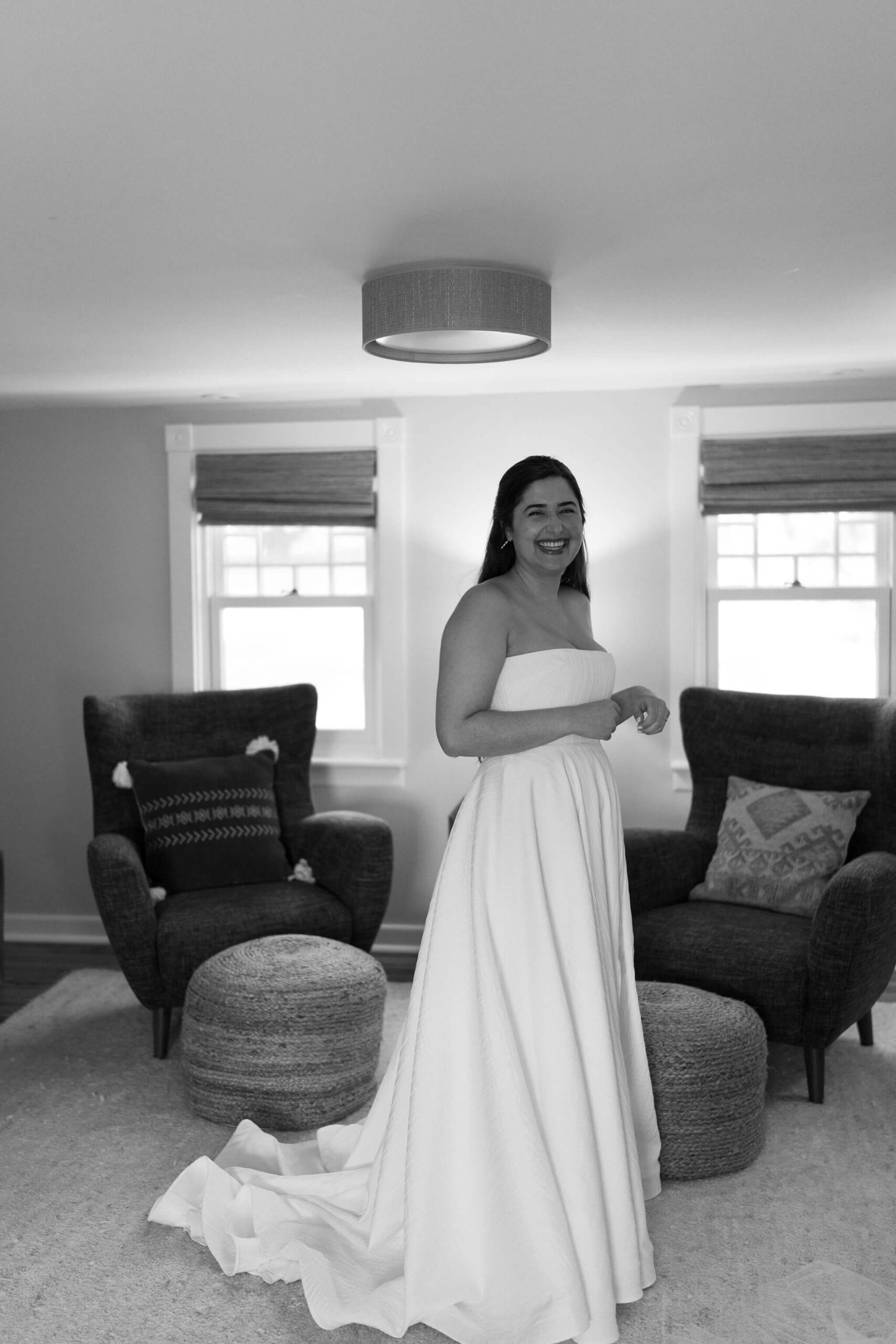 black and white image of a bride (long brown hair, strapless white wedding gown) standing in a bedroom laughing toward the camera