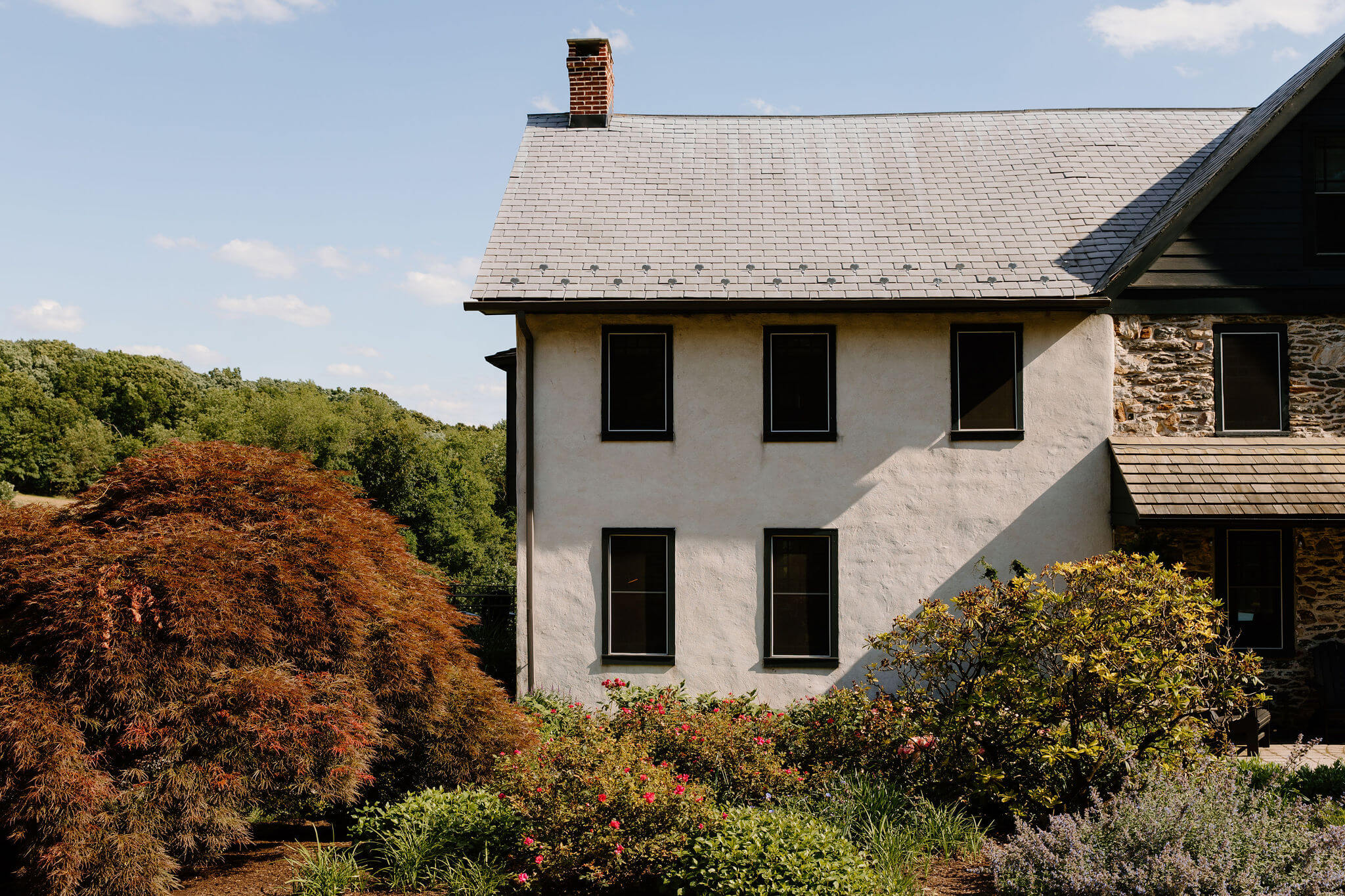 a stone farmhouse with a stucco addition, with pink rose bushes and other lush greenery and bushes in a garden in front of the house