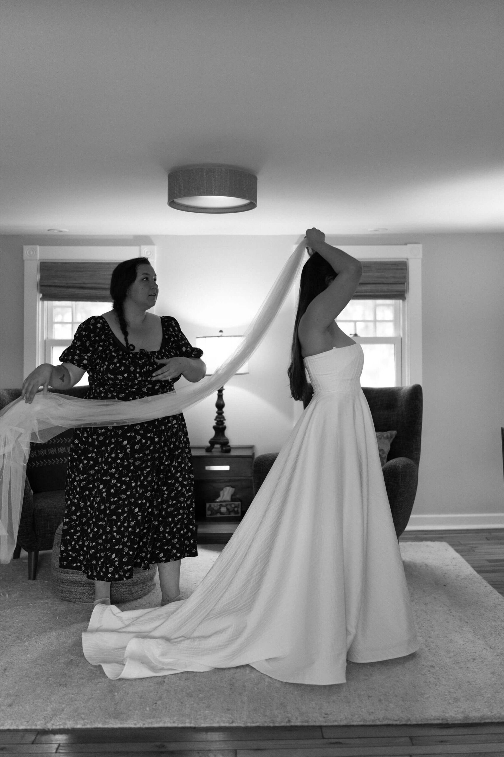 black and white image of bride's friend (black dress with floral accents) holding the end of a wedding veil while bride places it in her hair, wearing a long white bridal gown