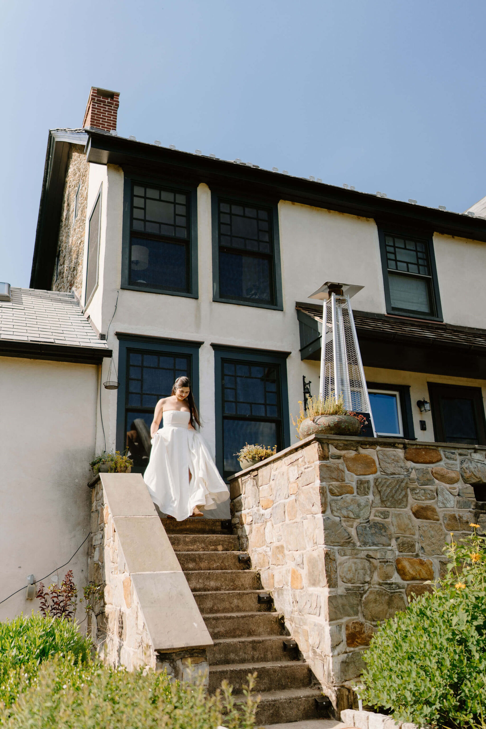 bride with brown hair walking down the stone stairs of a large farmhouse on a sunny day, holding the hem up of her strapless white wedding gown