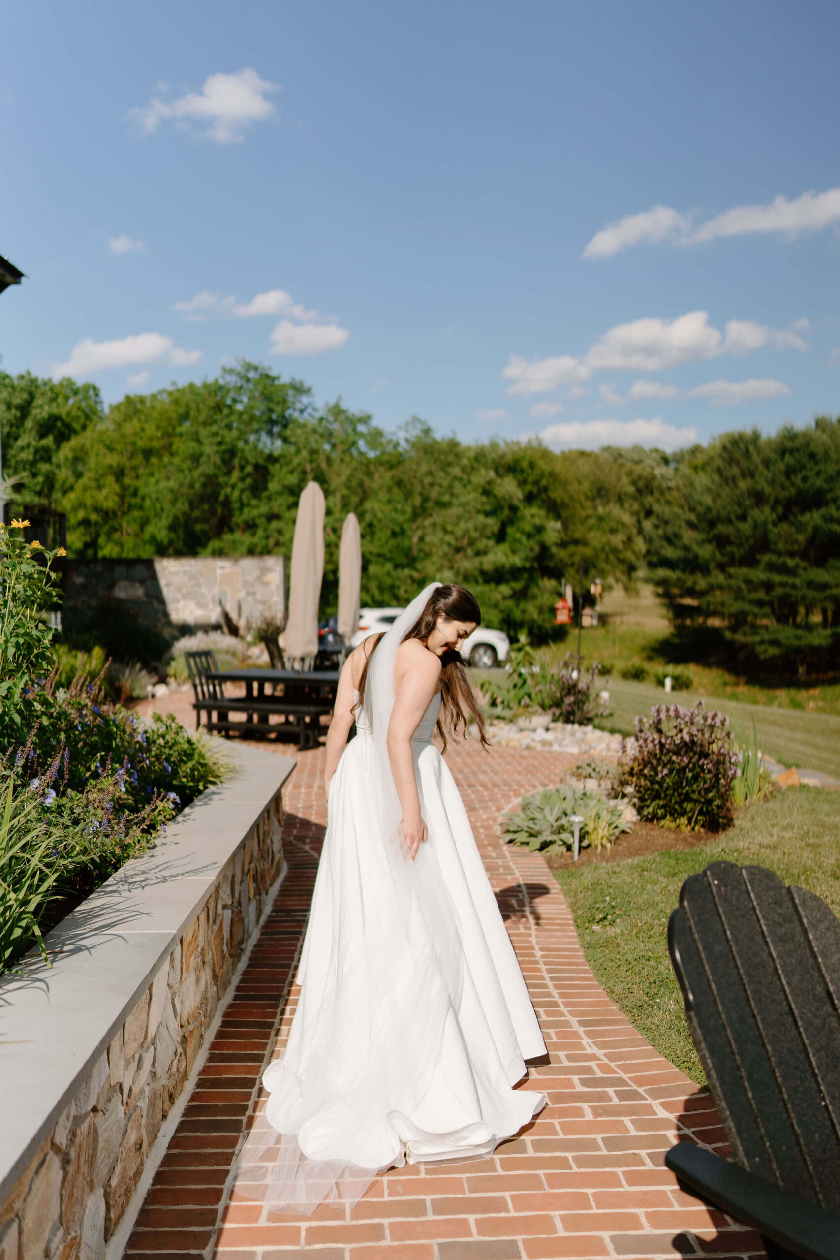 bride walking down a brick patio next to a stone wall, reaching behind her to straighten the train of her gown behind her