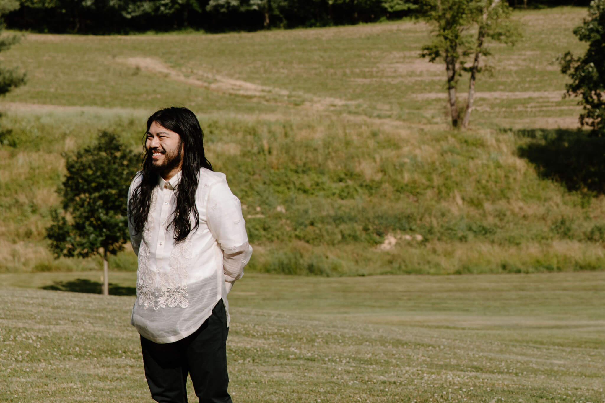 groom with long brown hair and a short beard, smiling off camera toward the bride, waiting for her to arrive to their wedding ceremony in a field