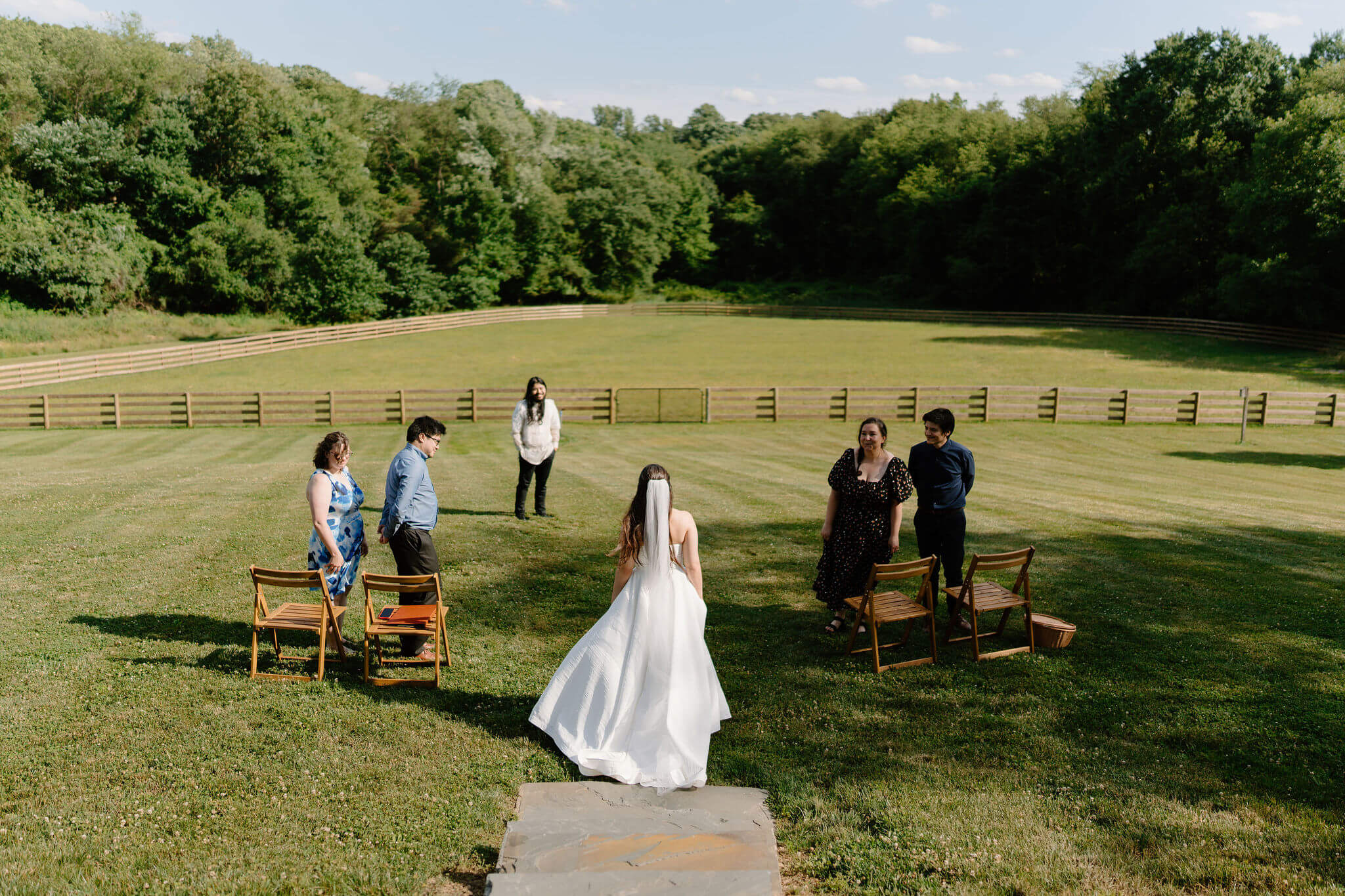 bride walking down a stone staircase into an open field, with two wooden chairs on either side and two friends on either side smiling at her, and the groom (white shirt, black pants) smiling at her