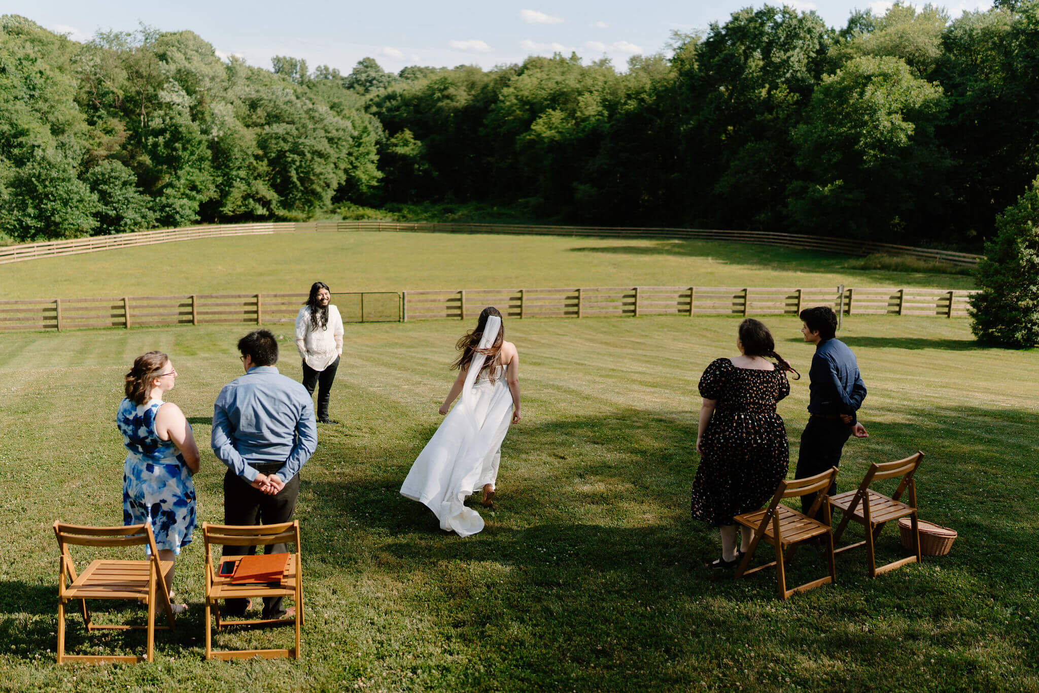 bride walking down a stone staircase into an open field, with two wooden chairs on either side and two friends on either side smiling at her, and the groom (white shirt, black pants) smiling at her