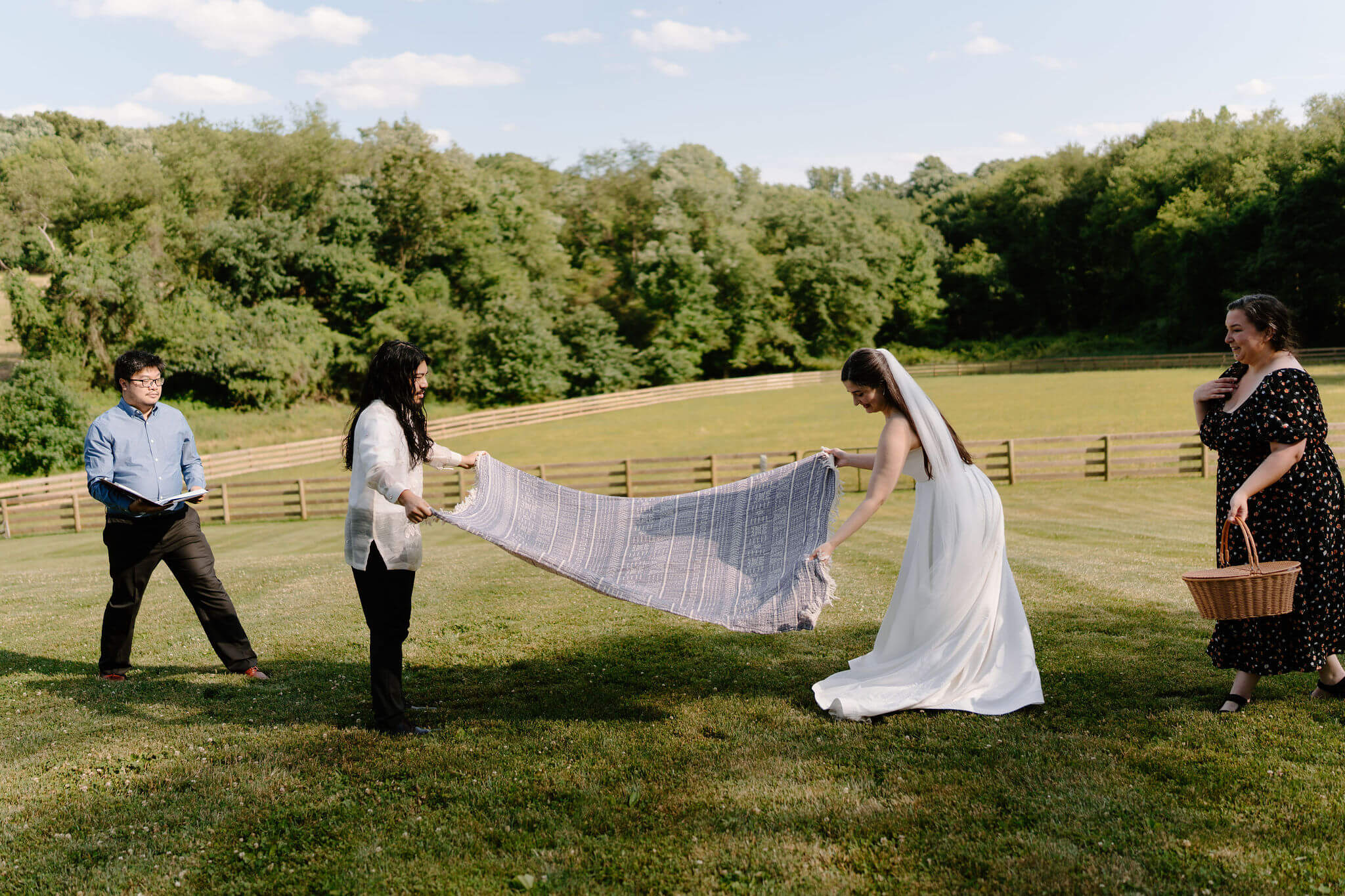 bride and groom spreading out a blanket in an open field for a portion of their wedding ceremony, while two friends stand next to them