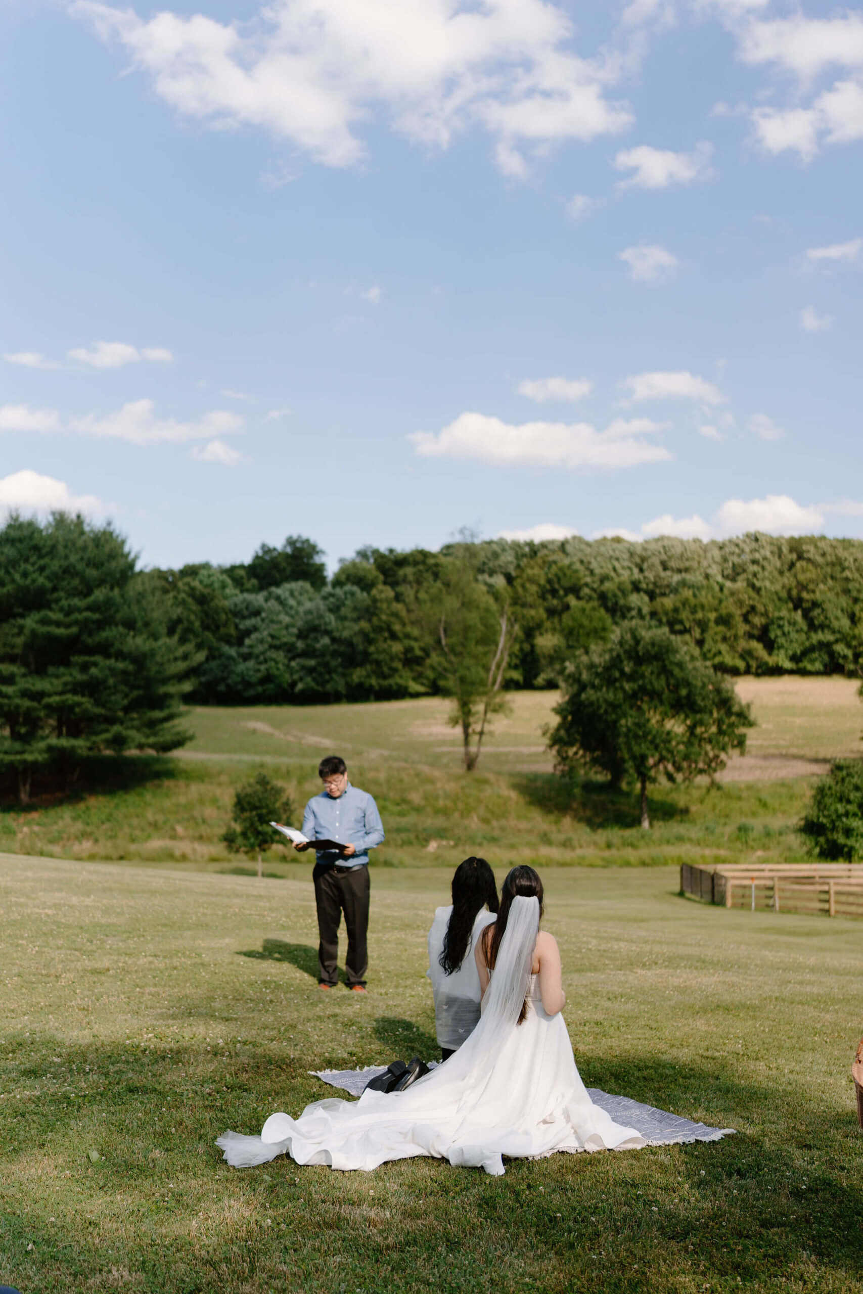 bride and groom kneeling on a blanket during their wedding ceremony, one guest standing next to them reading out of a binder