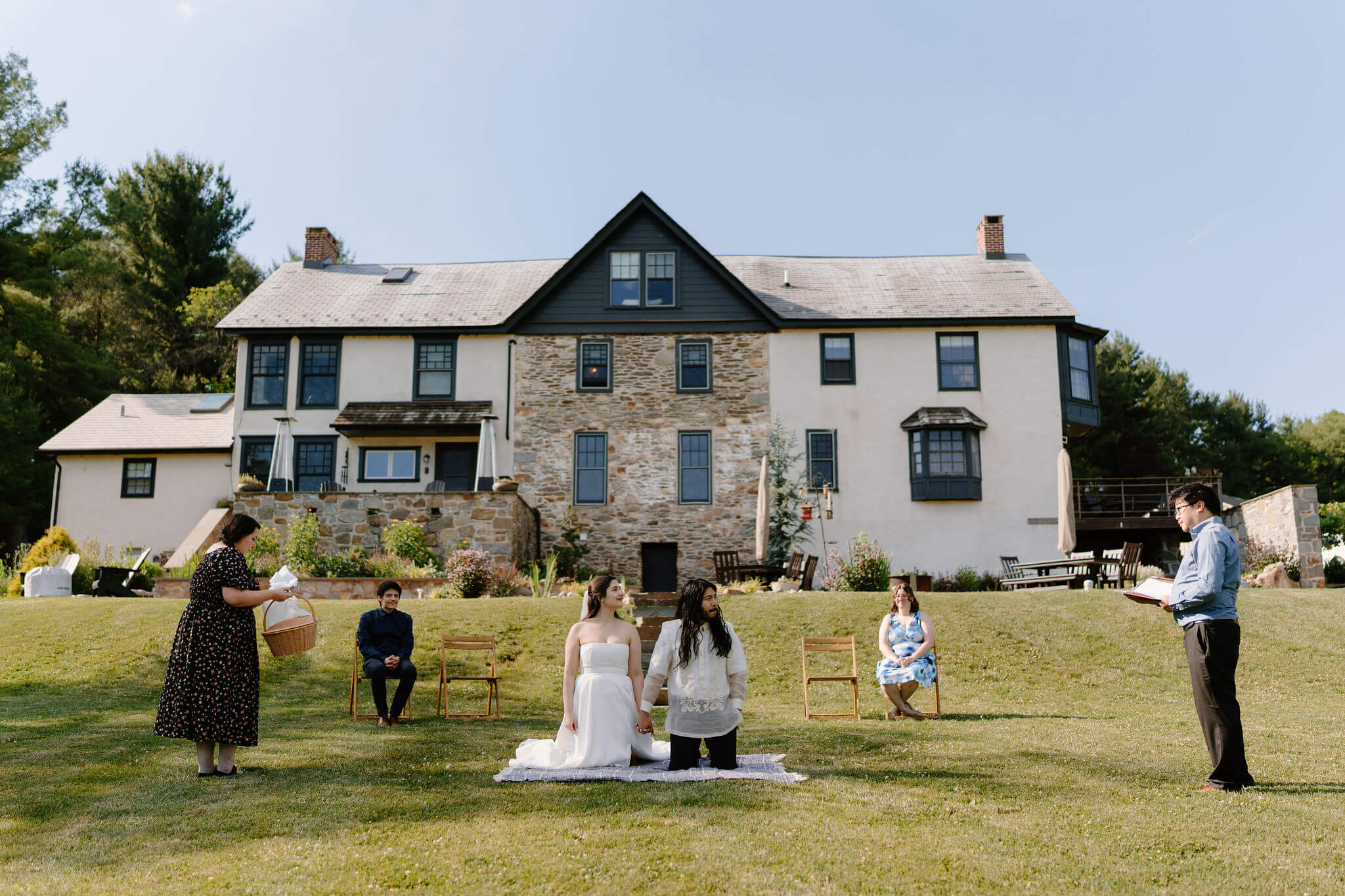 bride and groom kneeling on a blanket during their wedding ceremony, two guests seated behind them, with one guest standing on either side of them to perform a Filipino ceremony