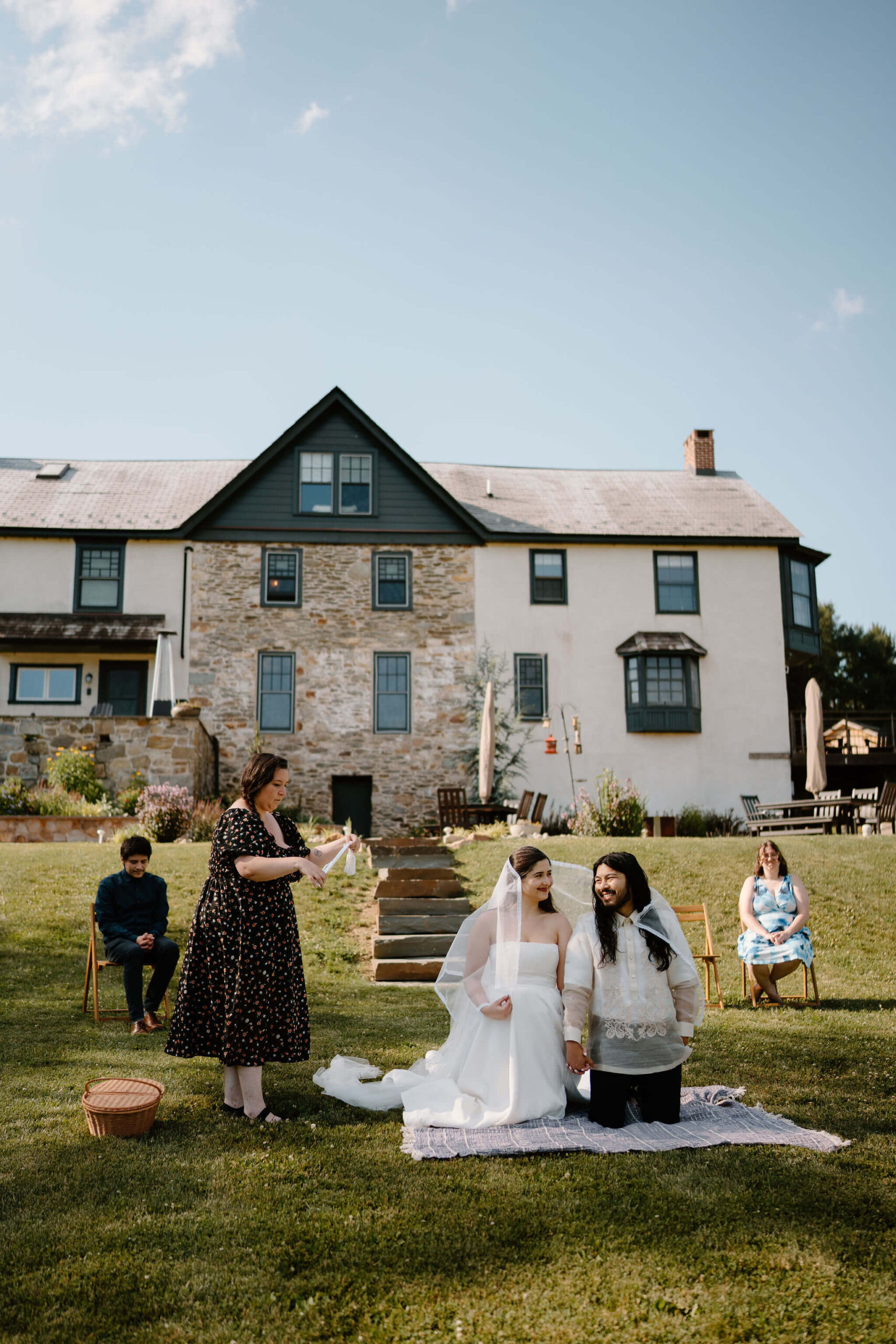 bride and groom kneeling on a blanket during their wedding ceremony, two guests seated behind them, with one guest standing next to them to perform a traditional Filipino lasso ceremony