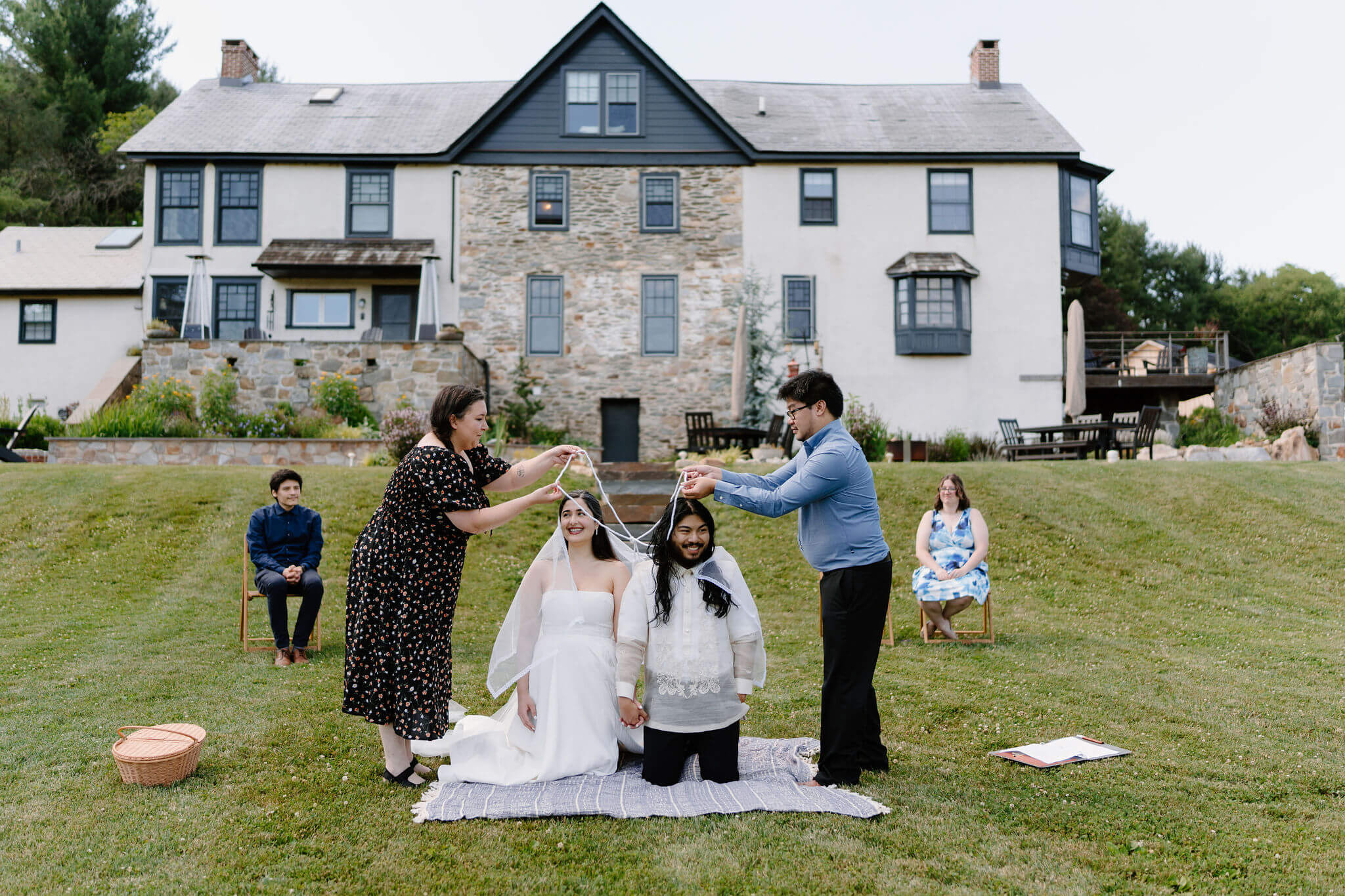 bride and groom kneeling on a blanket during their wedding ceremony, two guests seated behind them, with one guest standing on either side of them to perform a Filipino lasso ceremony