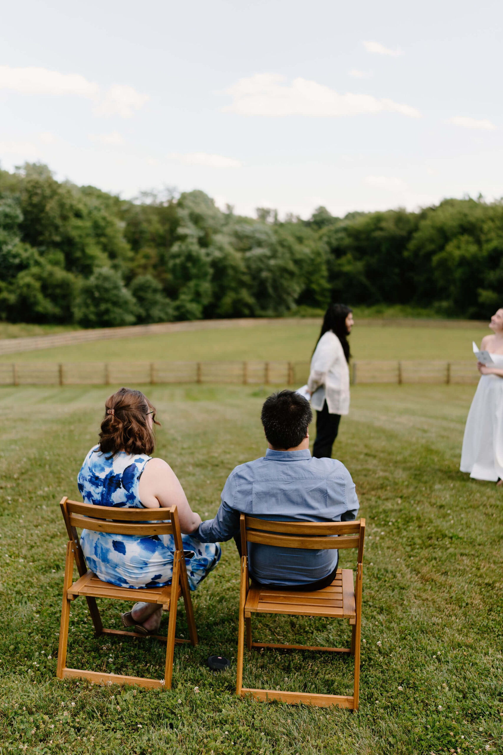 two guests seated and holding hands, watching their friends exchange wedding vows in an open field