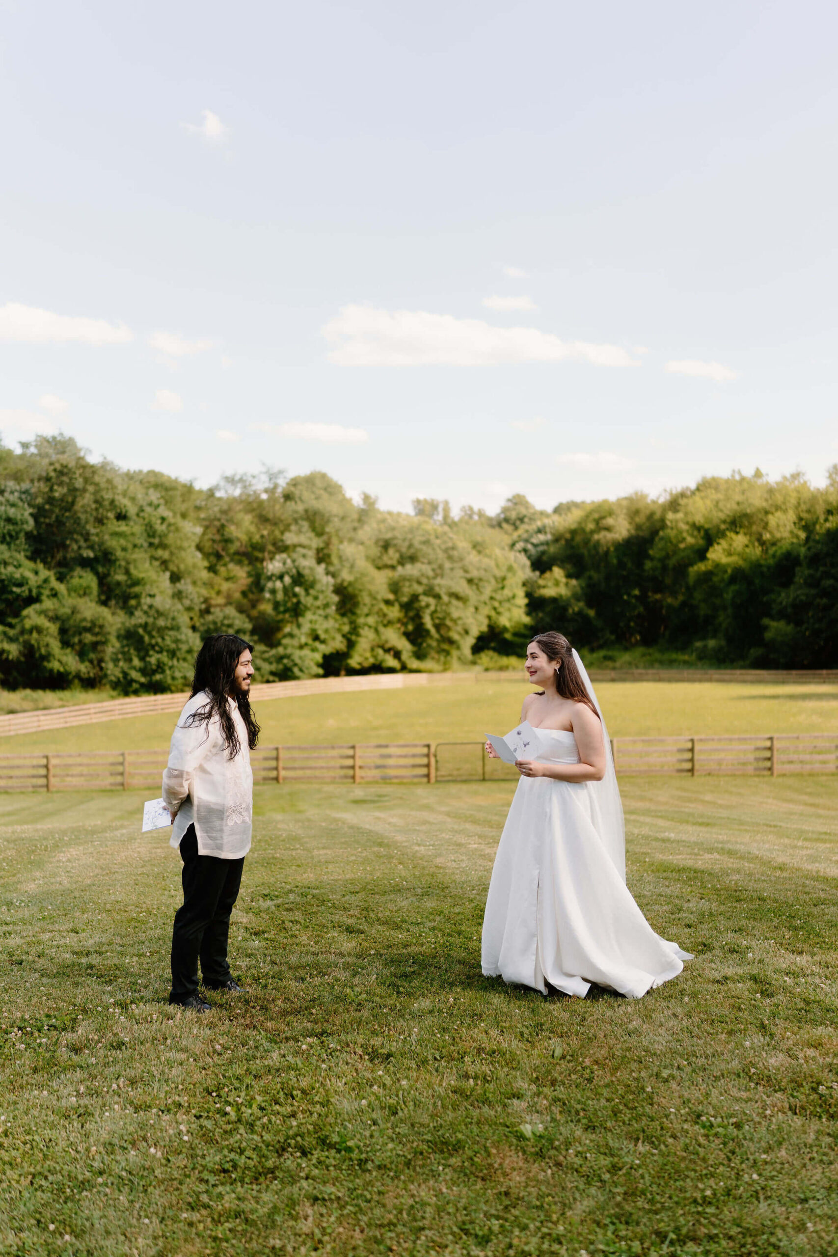 bride and groom standing in an open field on a sunny day, facing each other and exchanging vows