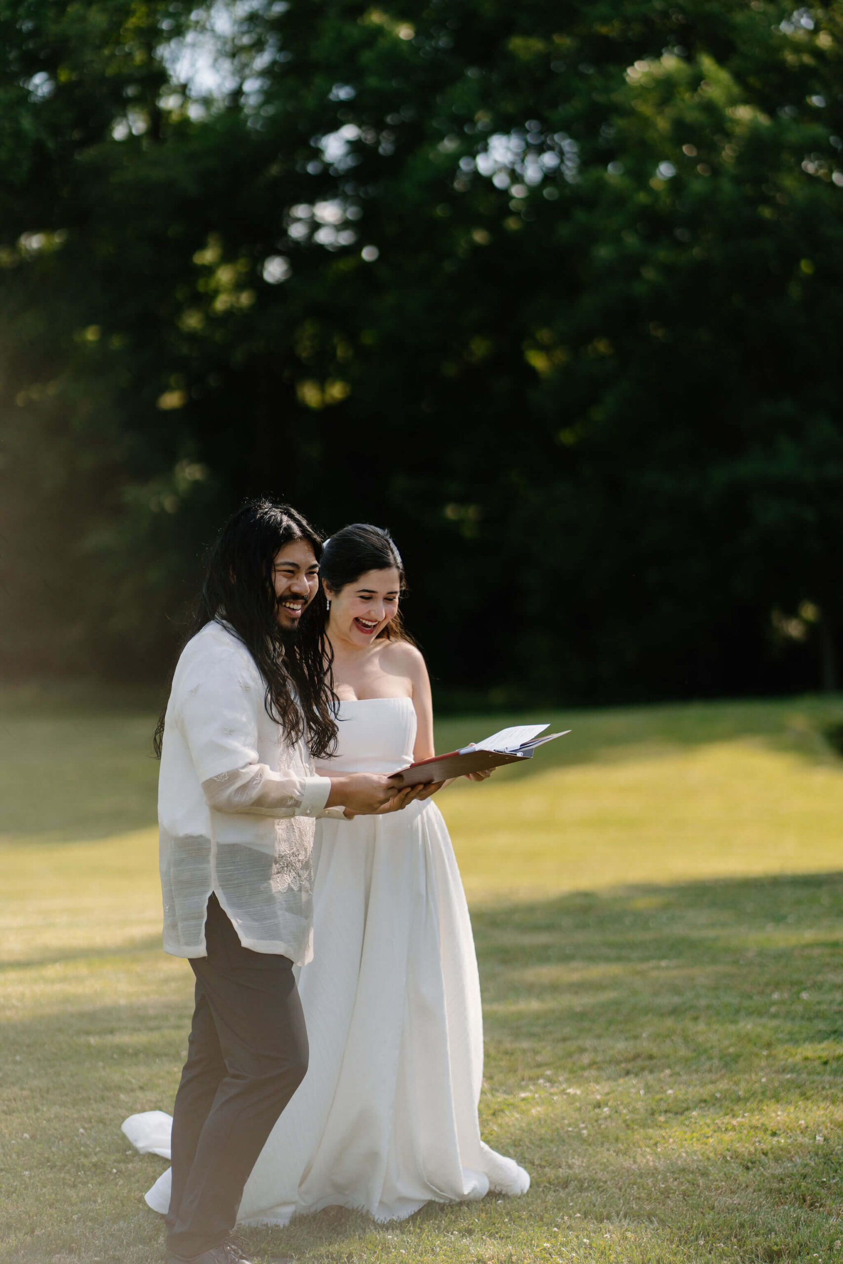 bride and groom standing in an open field on a sunny day, reading out of a binder for their wedding ceremony and laughing toward their guests