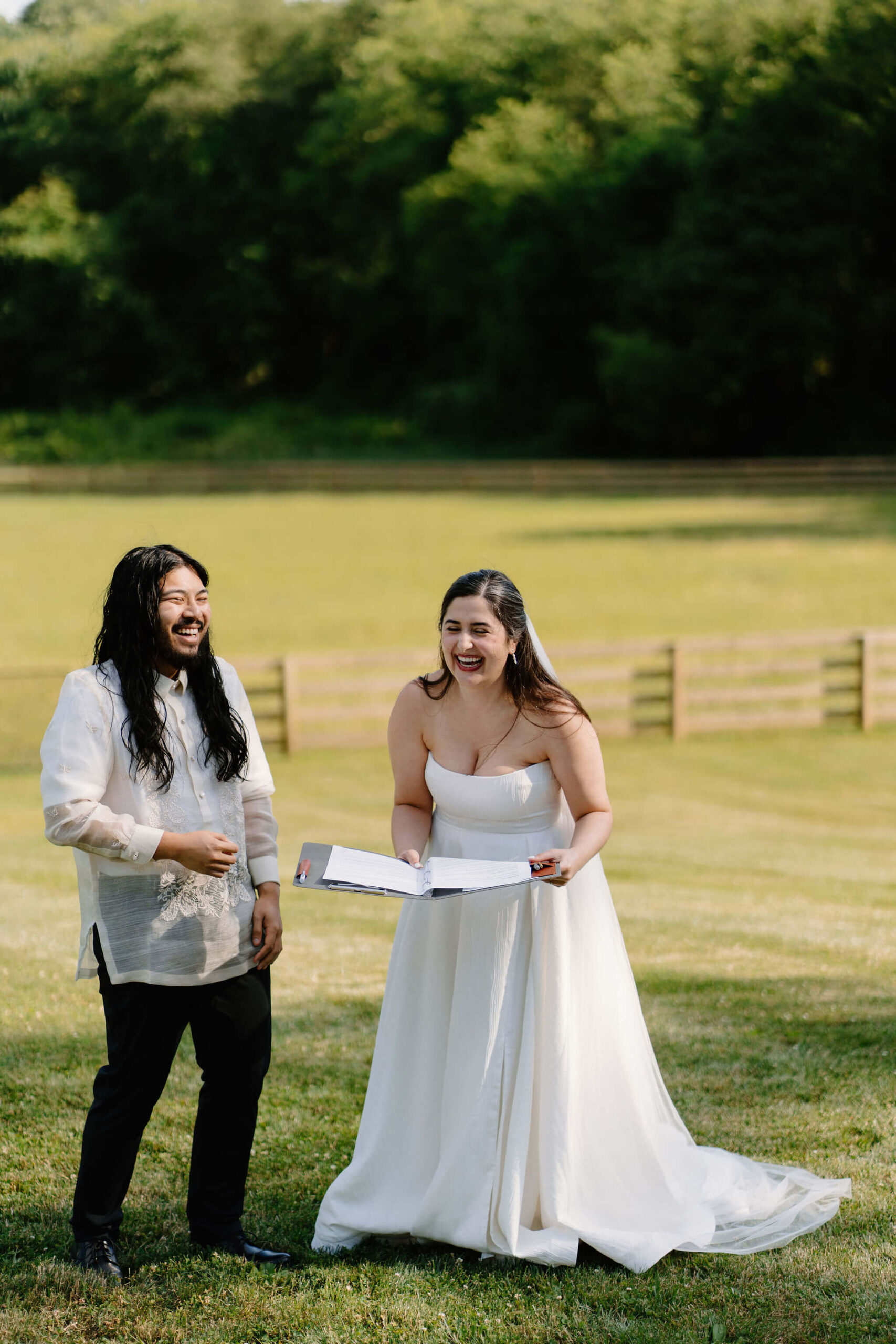 bride and groom standing in an open field on a sunny day, reading out of a binder for their wedding ceremony and laughing toward their guests