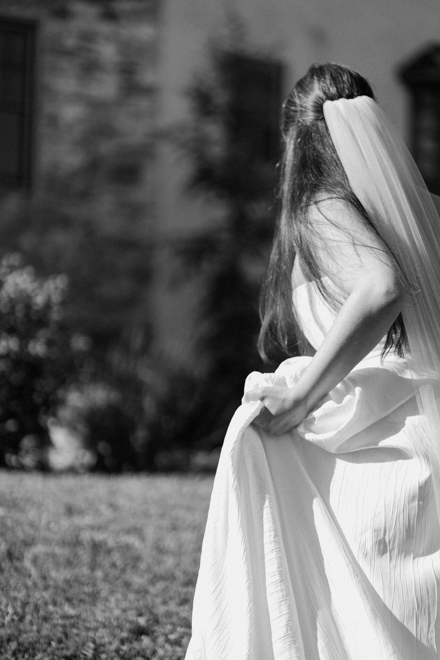 black and white image of bride walking up a hill, hiking up the hem of her wedding dress