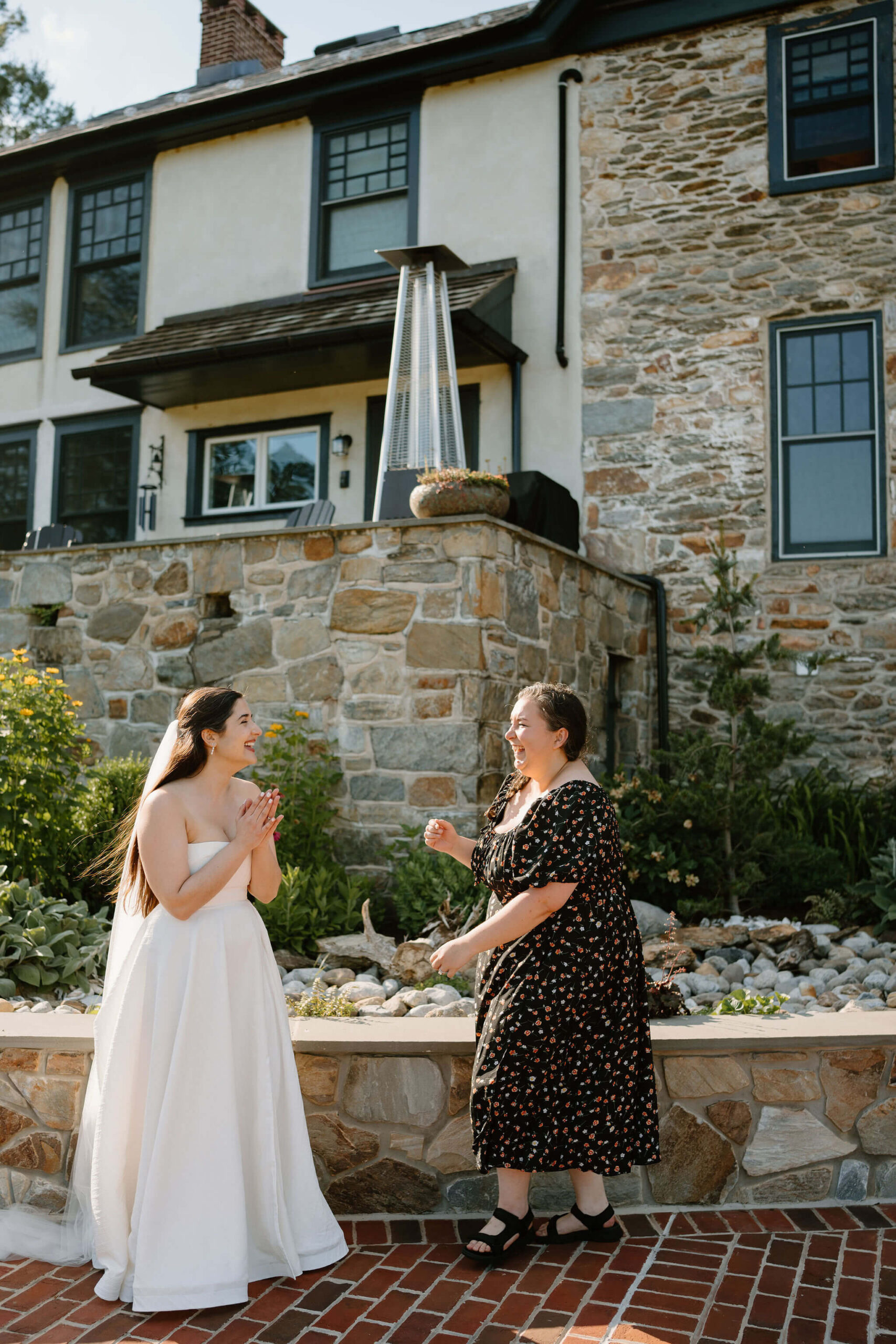 bride and her friend laughing loudly toward each other in front of a large stone farmhouse