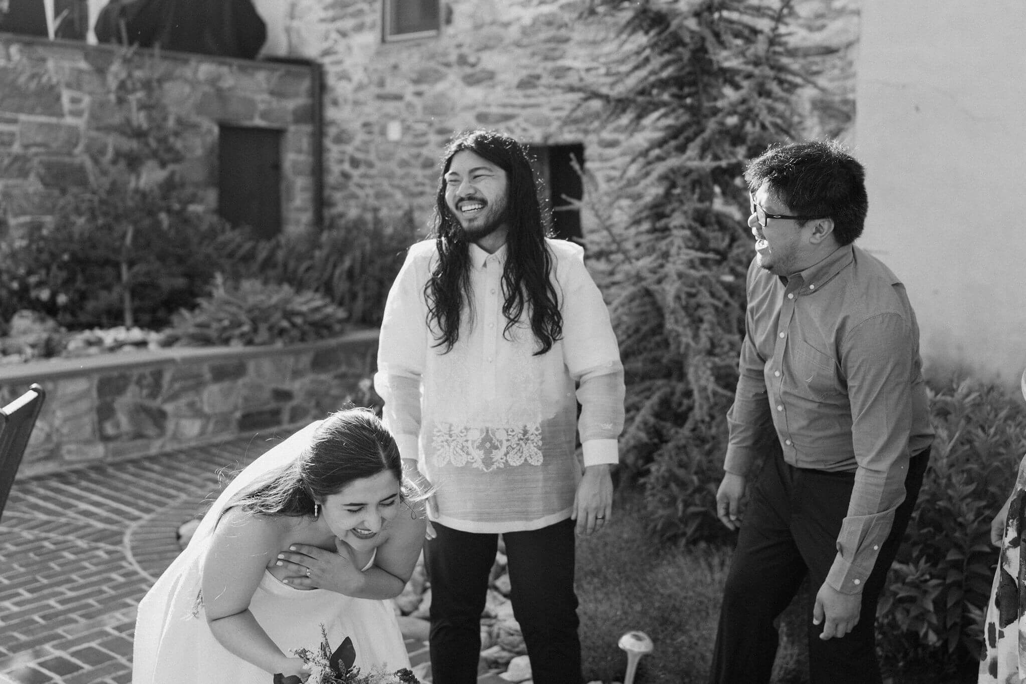 black and white image of bride, groom, and friend laughing together, bride doubled over and holding her chest