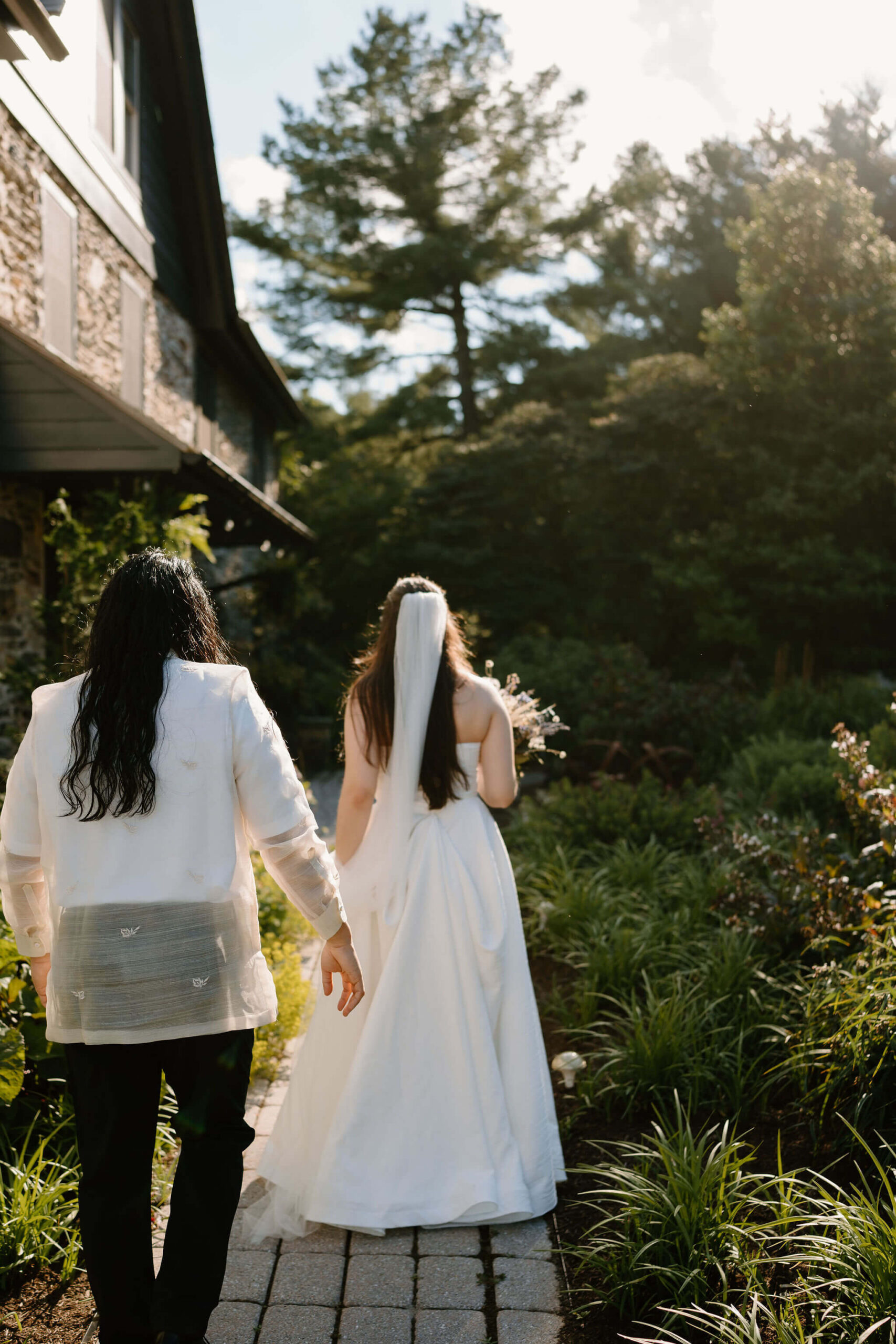 bride and groom walking up a stone garden path next to a large stone farmhouse. away from the camera, sun backlighting them