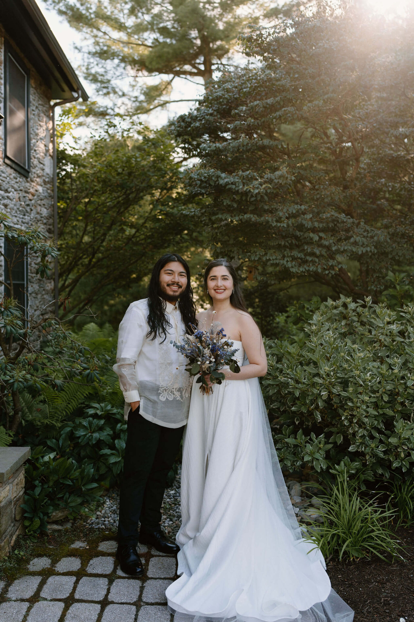 bride (white strapless gown, long brown hair, floor length veil) holding flowers, and smiling at the camera next to groom (white embroidered shirt, black pants, long brown hair) smiling toward the camera, on a stone path in front of a lush green garden with trees