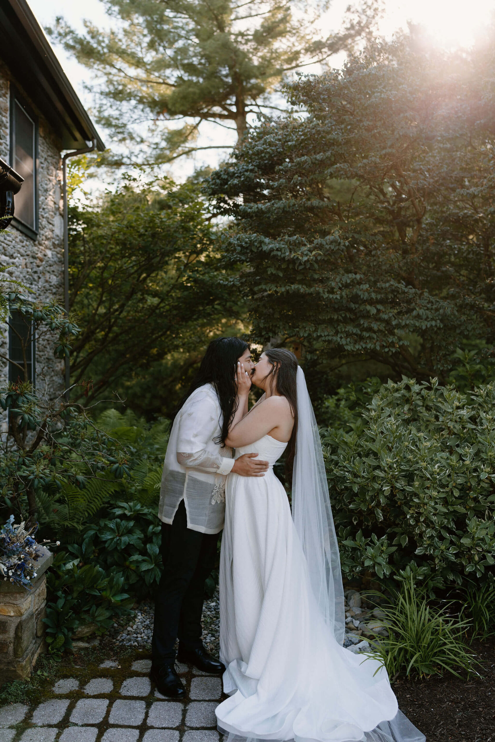 bride (white strapless gown, long brown hair, floor length veil) holding groom's face and kissing him (white embroidered shirt, black pants, long brown hair) on a stone path in front of a lush green garden with trees