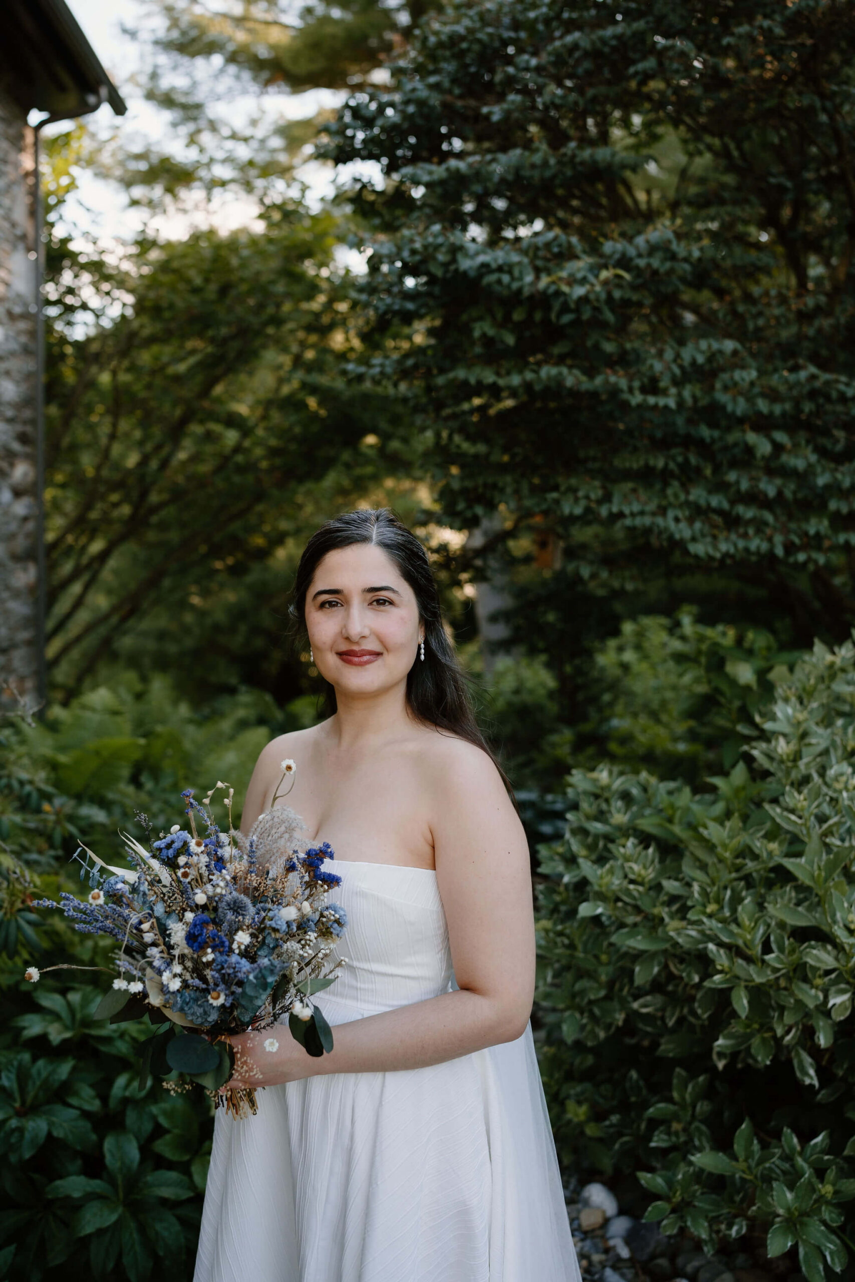 bride smiling softly toward the camera, long brown hair over her shoulders, holding dried flower bouquet of blue and purple flowers against a lush green background