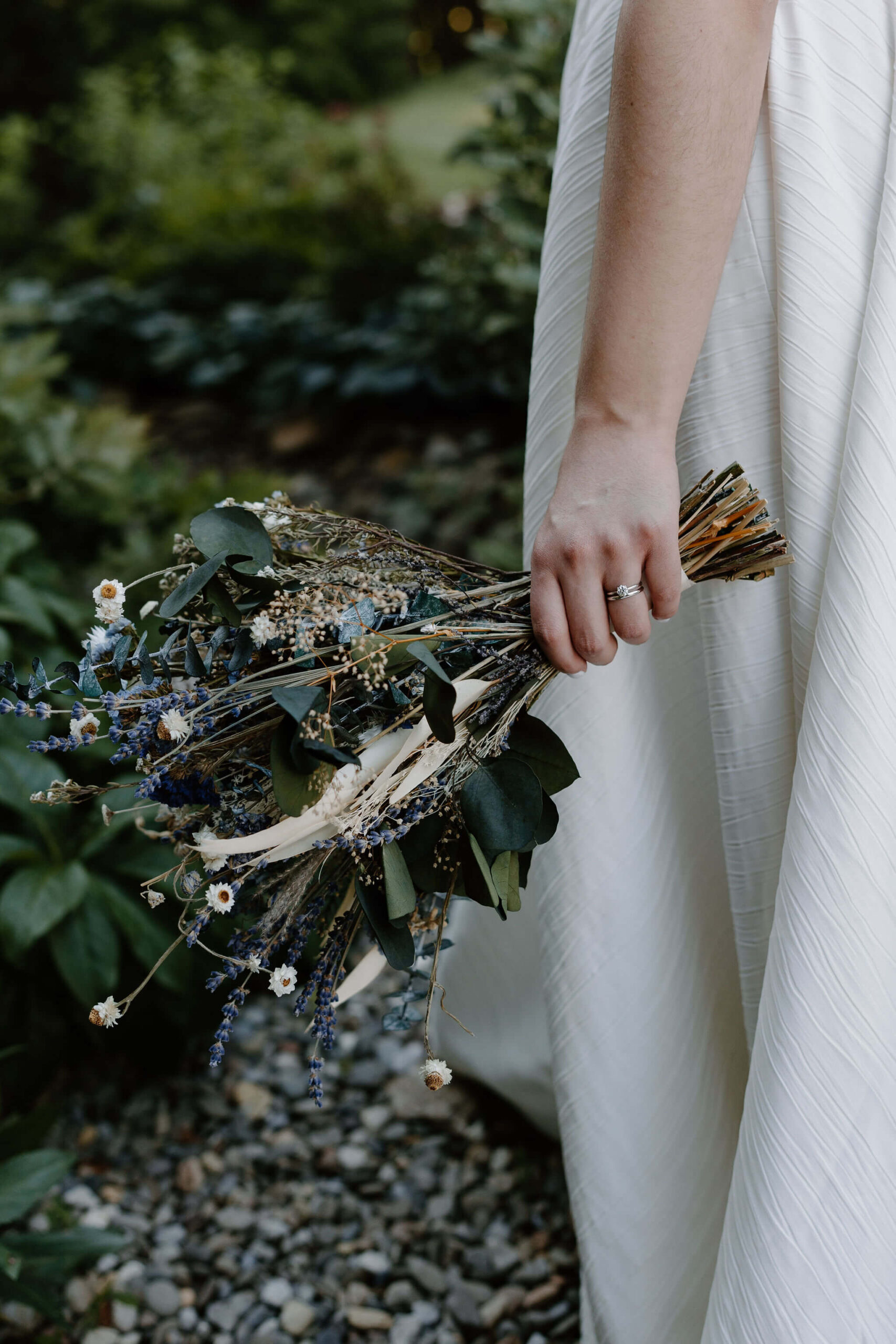 close up of bride's hand holding her bouquet of dried blue and purple flowers, with her textured dress in the background