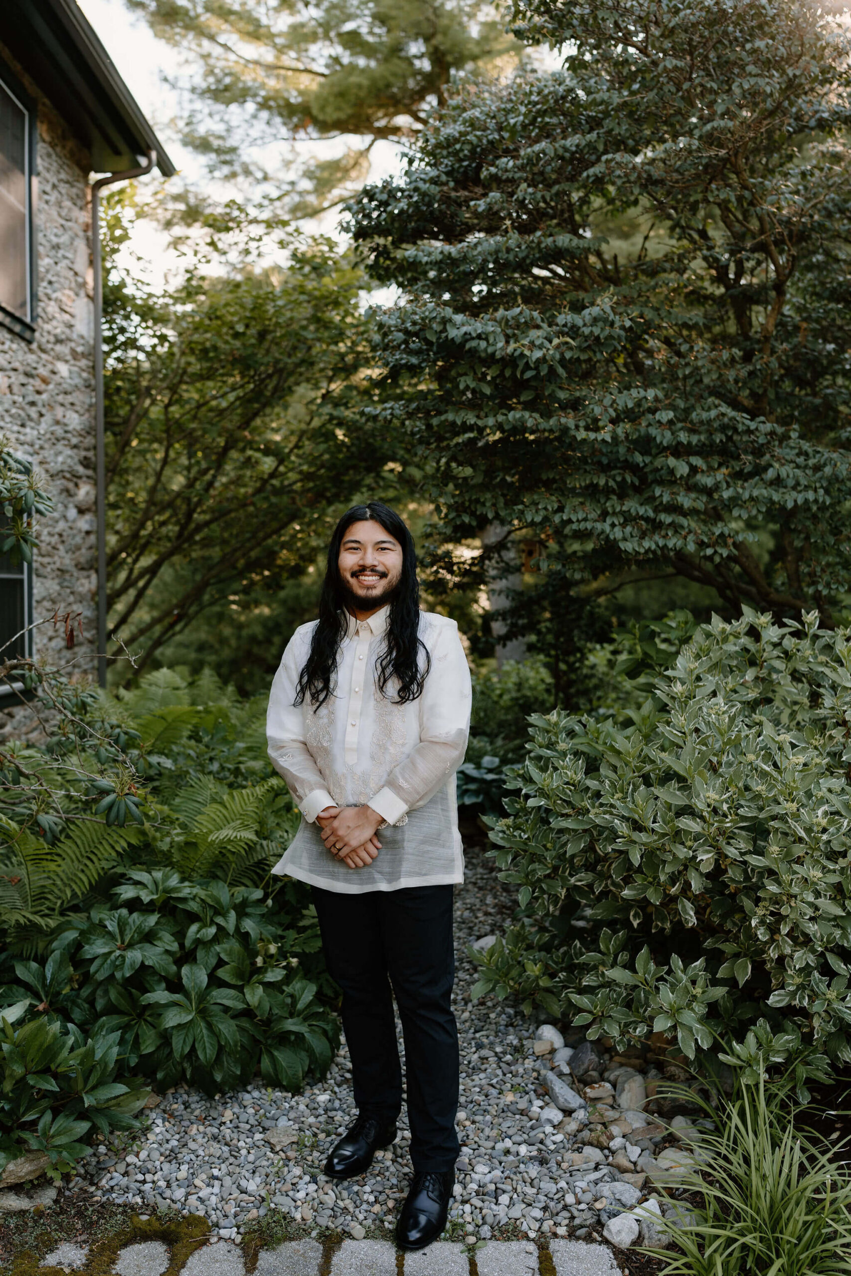 groom (white embroidered shirt and black pants, with long brown hair and short beard) smiling with hands crossed in front of him, on a stone path with a lush green garden behind him