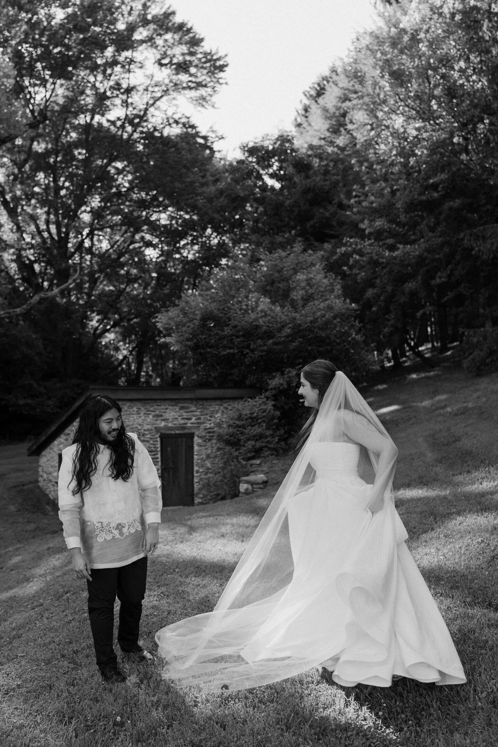 black and white image of bride and groom next to a pond, surrounded by tall green trees, dancing and smiling at each other while bride swishes her train around