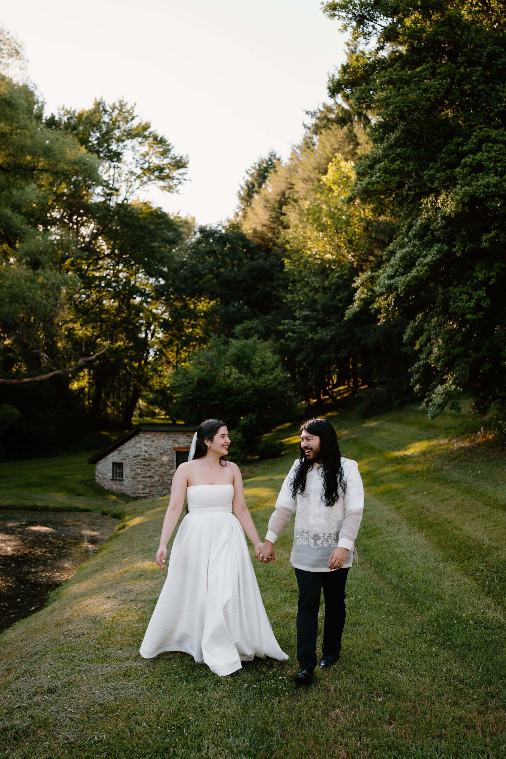 bride and groom next to a pond, surrounded by tall green trees, holding hands and walking toward the camera while smiling at each other