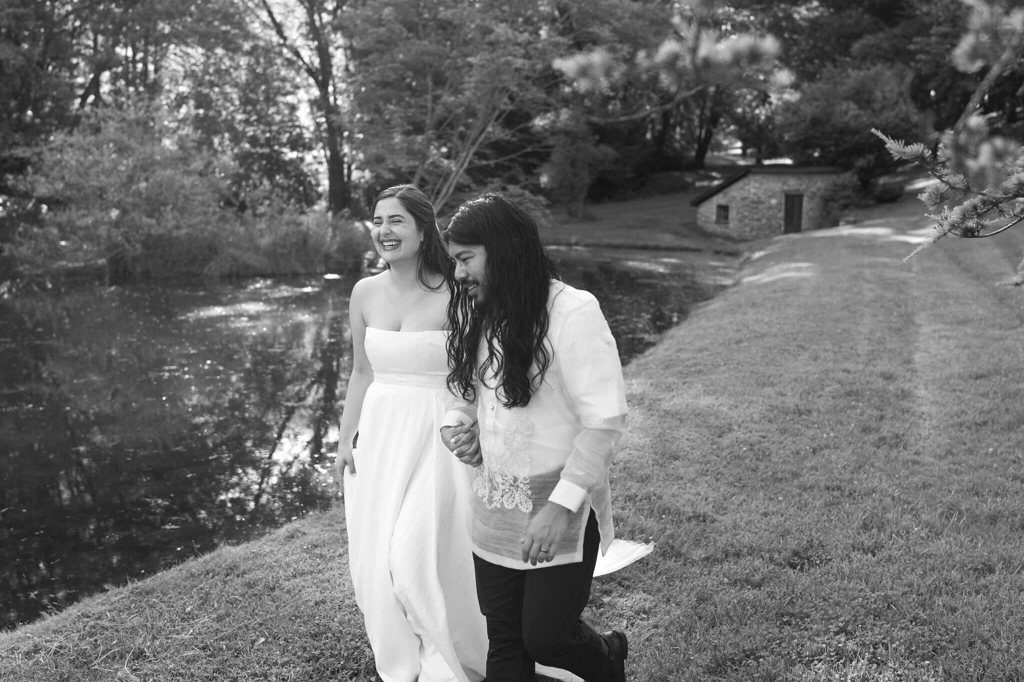 black and white image of bride and groom next to a pond, surrounded by tall trees, holding hands and walking toward the camera while laughing toward each other