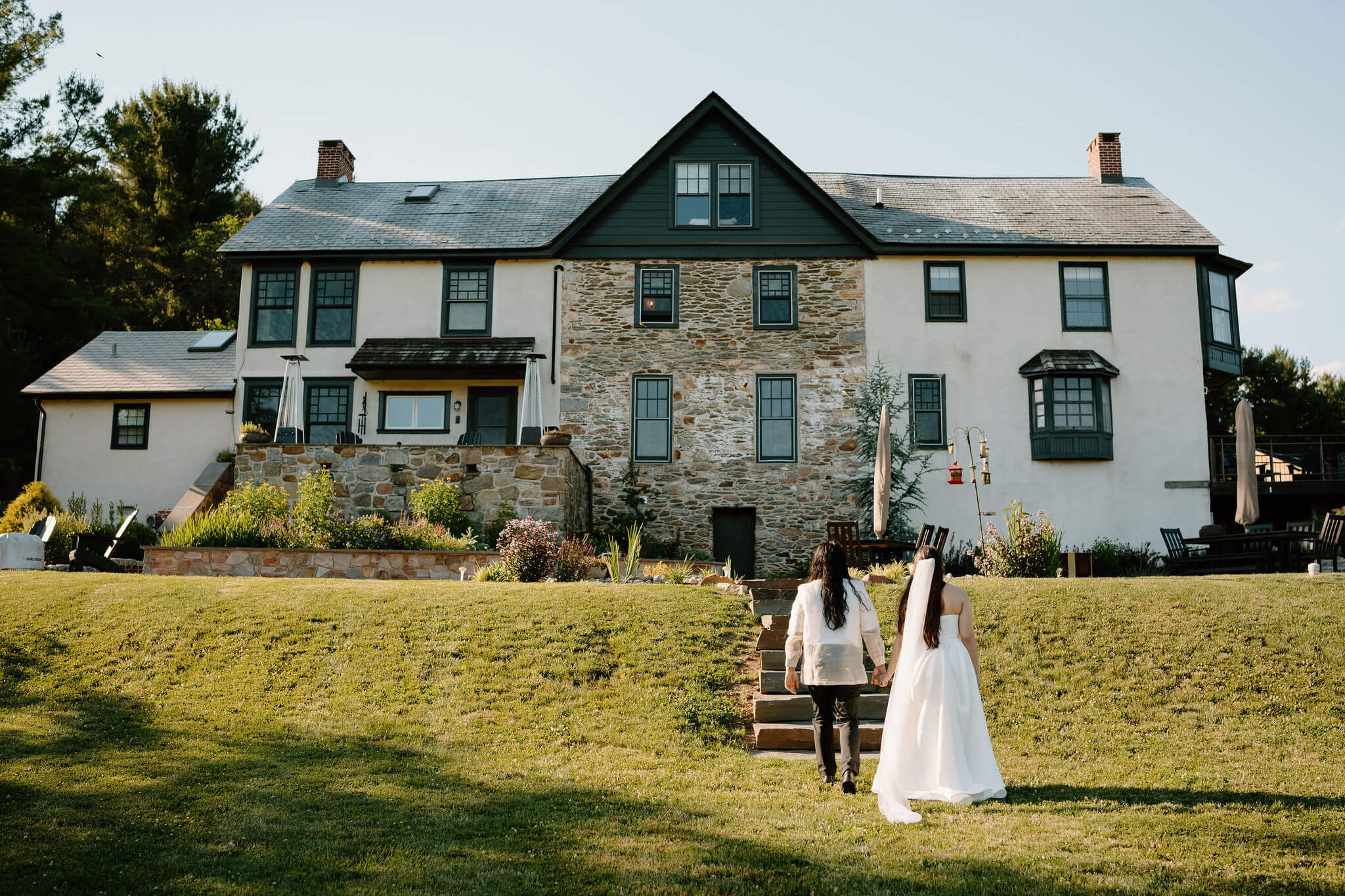 bride and groom holding hands and walking up a stone path toward a large stone farm house, away from camera