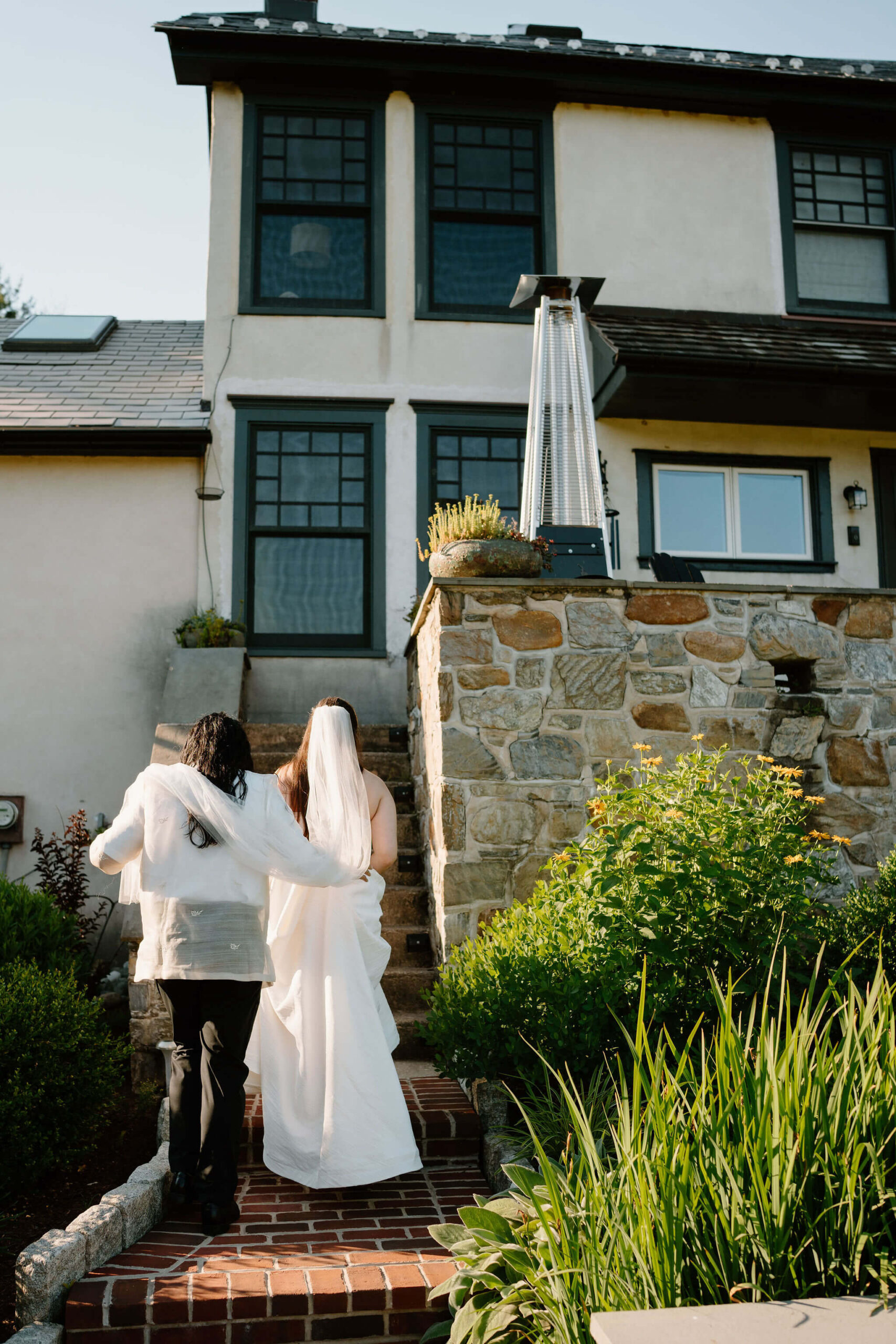 groom holding bride's veil over his shoulders while she walks up stairs to a stone patio