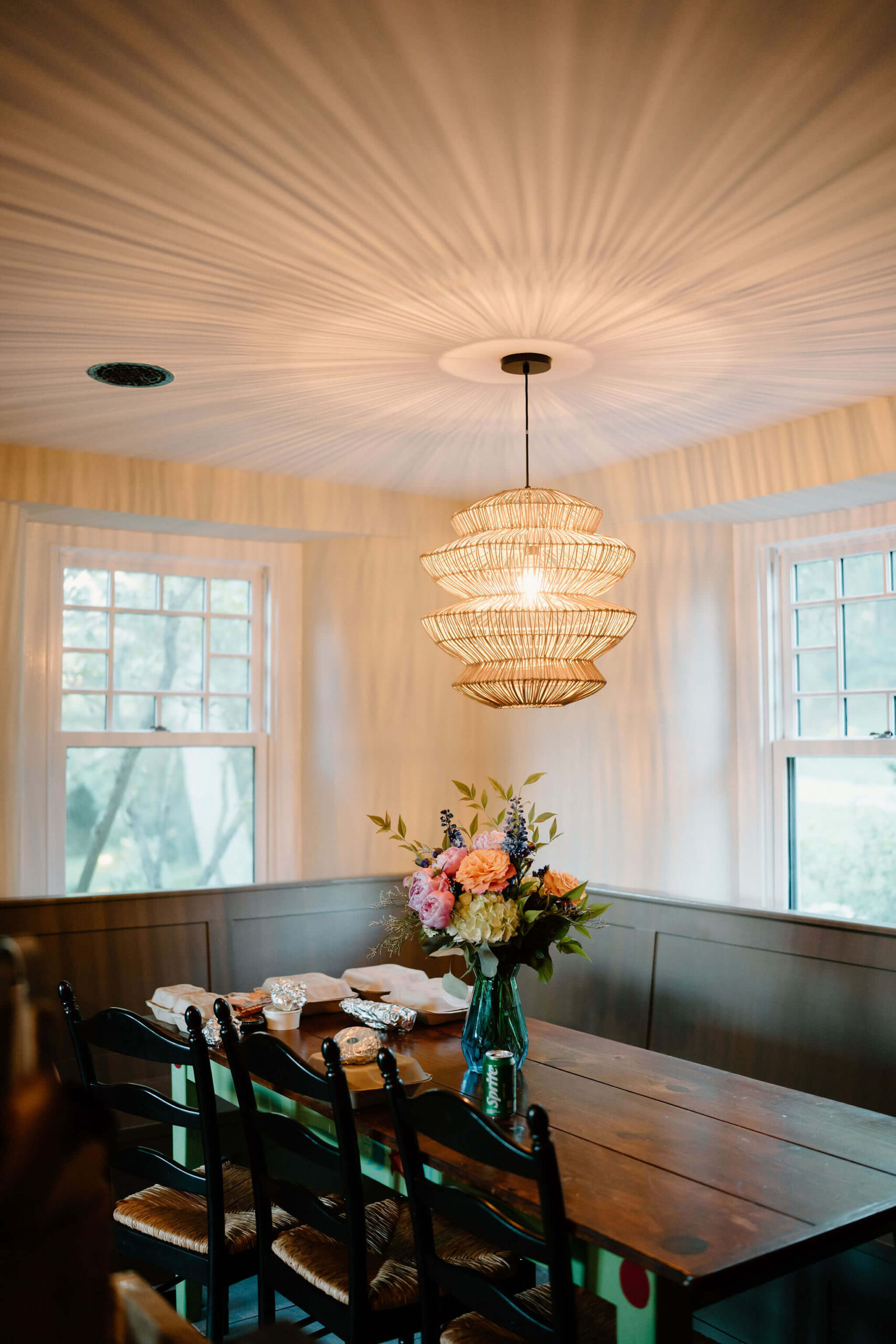 flowers on a wooden kitchen table with chairs, takeout containers for dinner scattered on it, with a large light fixture throwing interesting shadows on the ceiling