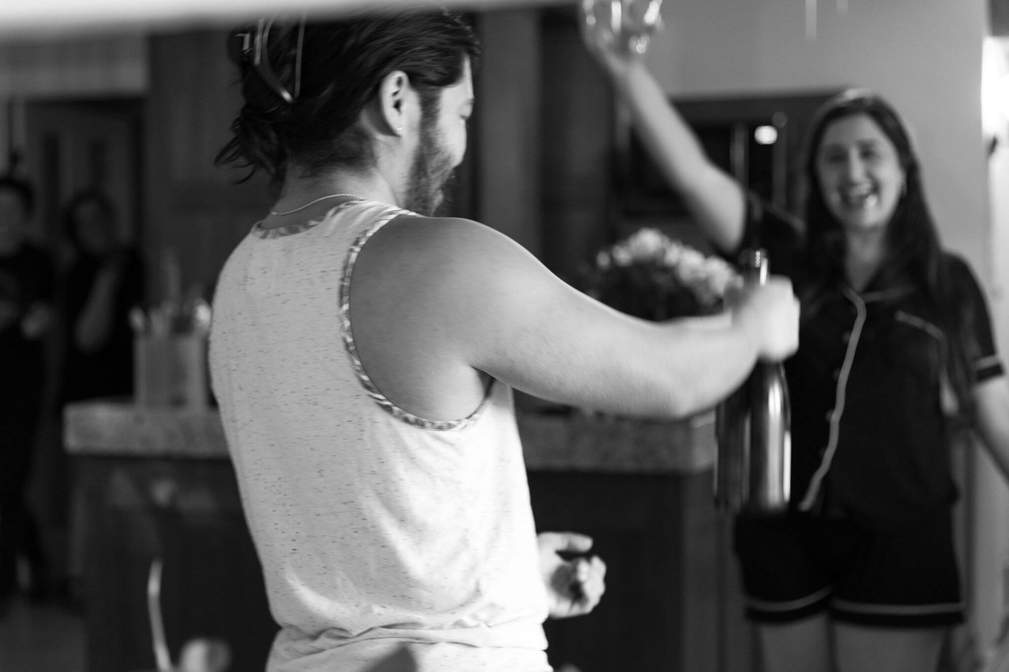 black and white image of groom (in pajamas) just opening a bottle of wine, while bride (also in pajamas) cheers in the background