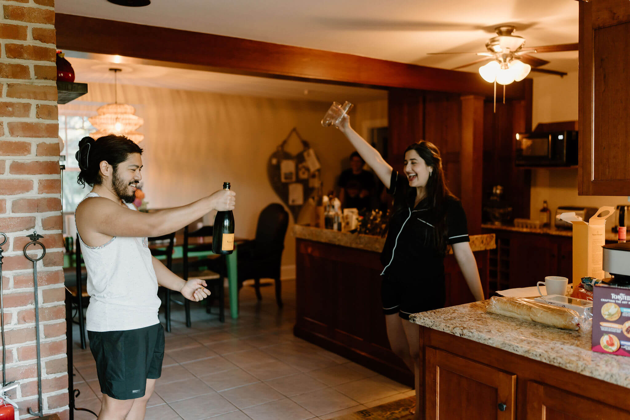 groom (in pajamas) opening a bottle of champagne, while bride (also in pajamas) cheers in the background