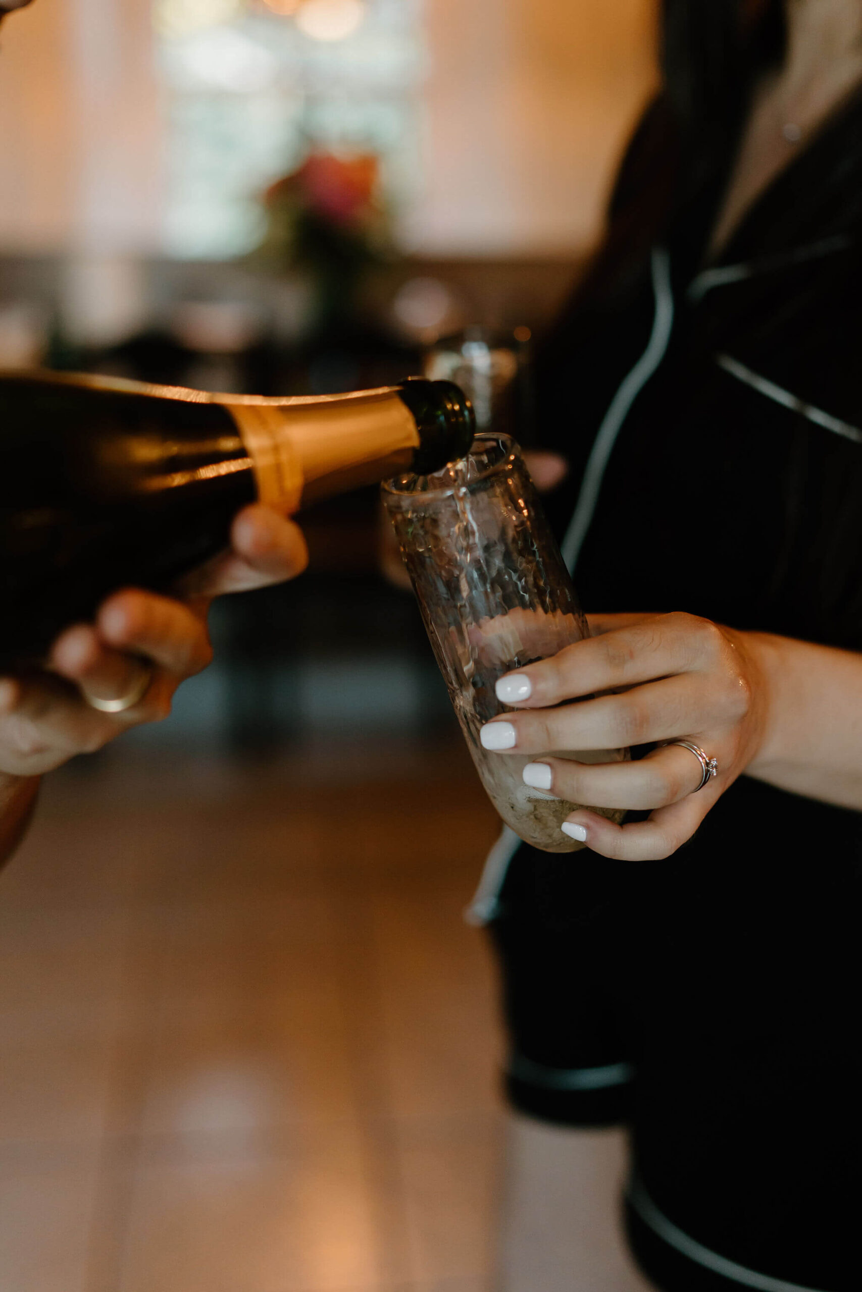 groom pouring champagne into a stemless flute held by the bride, focusing on her engagement rings