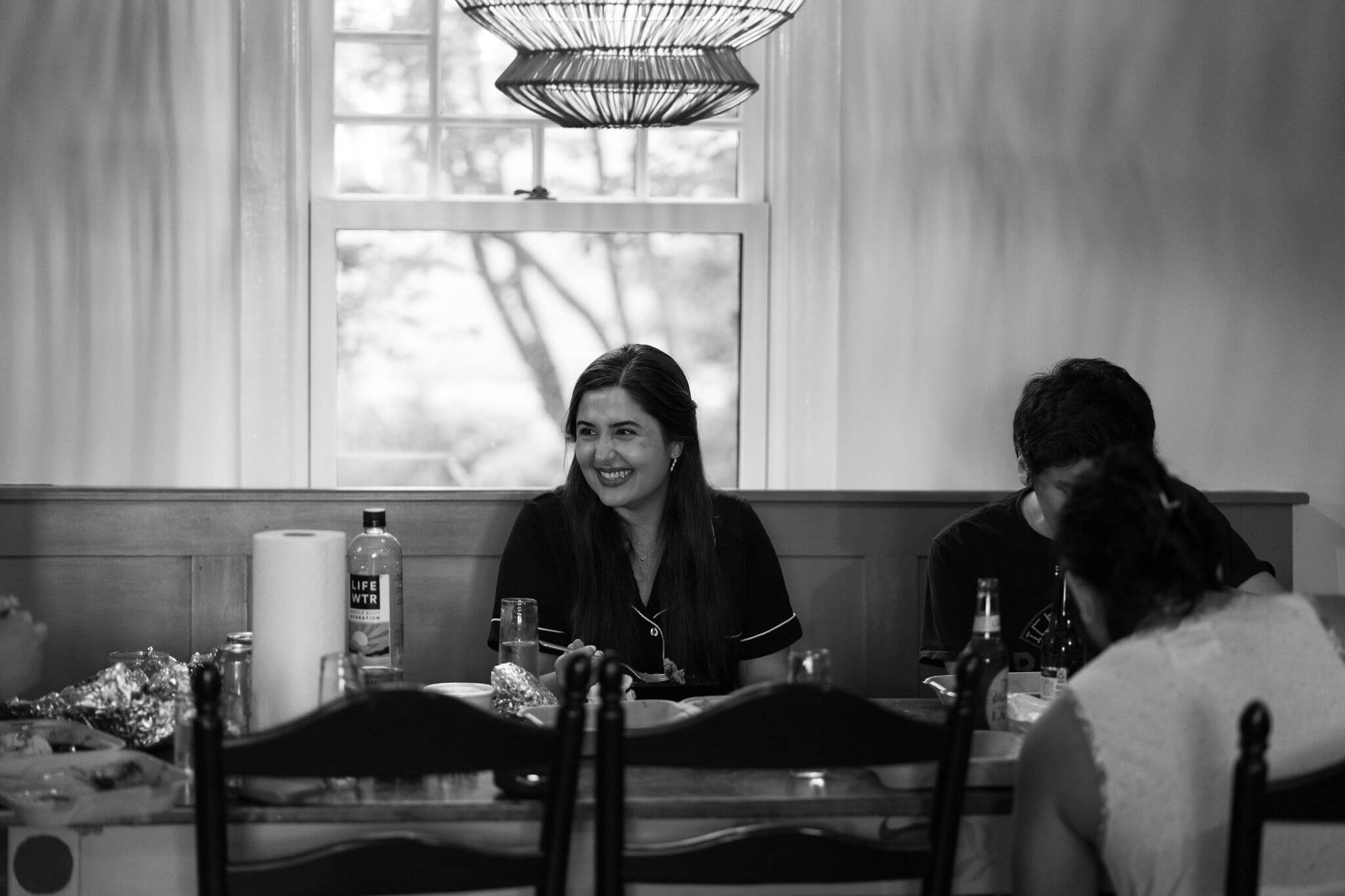 black and white image of bride, wearing pajamas and sitting at kitchen table having dinner, laughing off screen