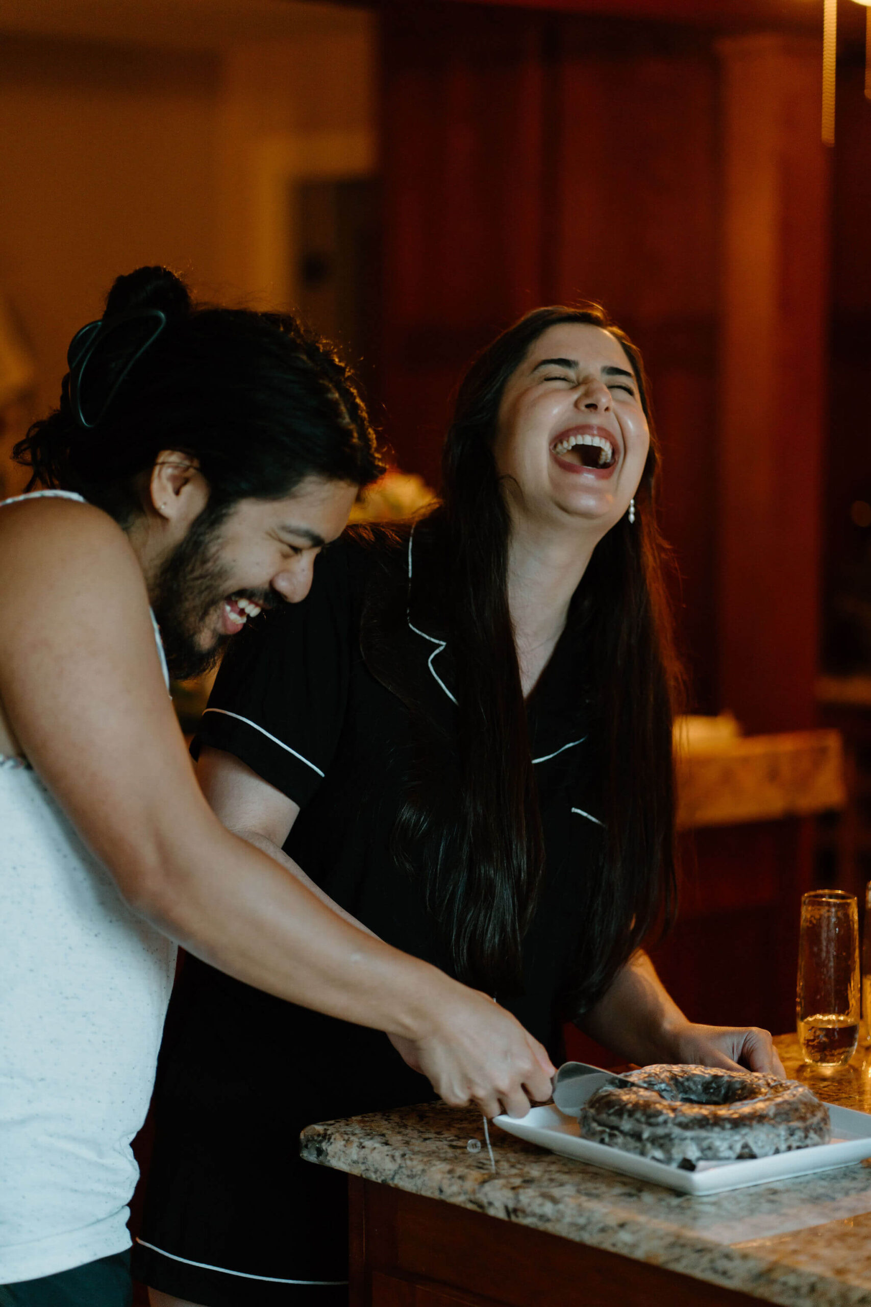 bride and groom (both in pajamas) laughing while cutting a large chocolate doughnut in lieu of a wedding cake