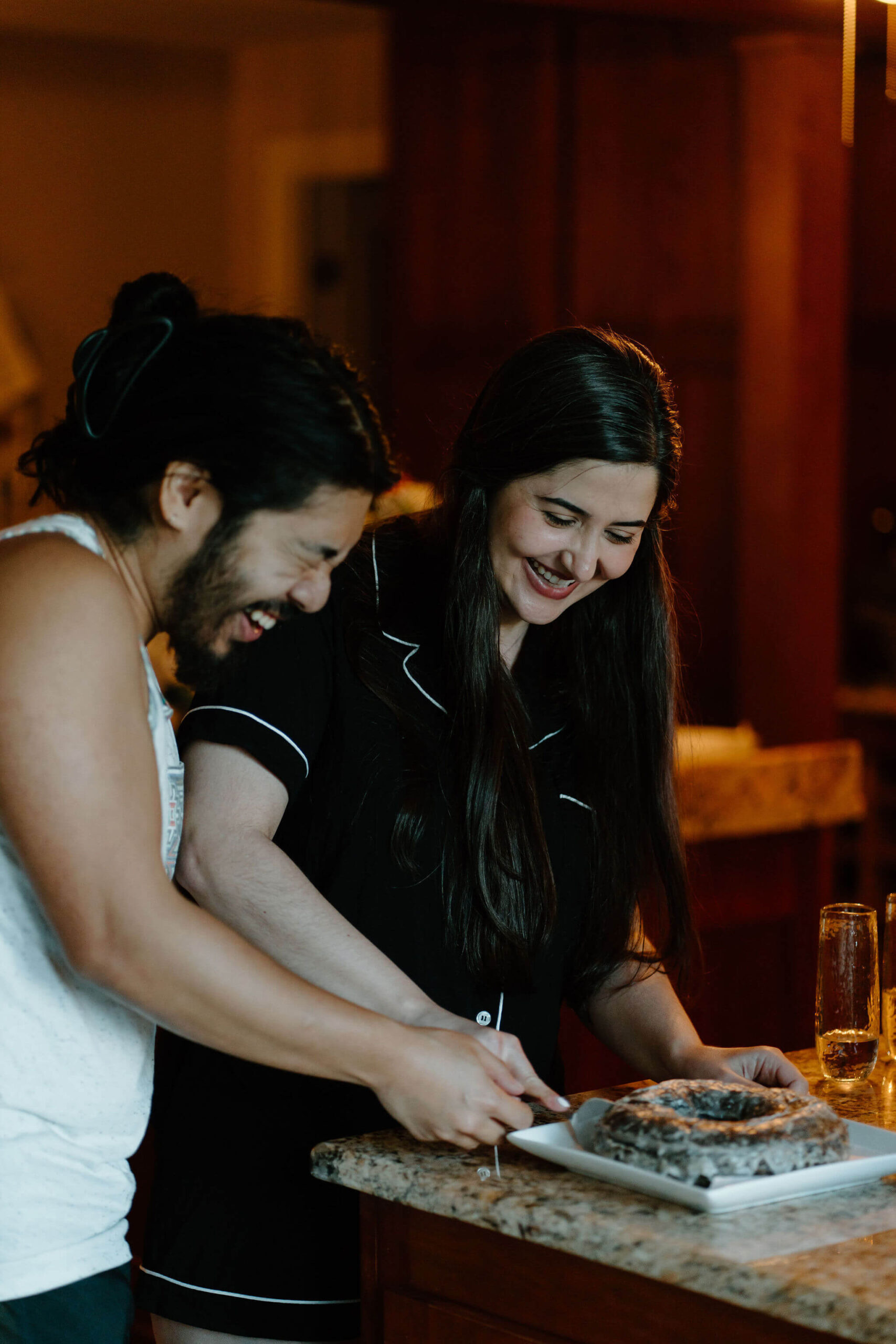 bride and groom (both in pajamas) laughing while cutting a large chocolate doughnut in lieu of a wedding cake