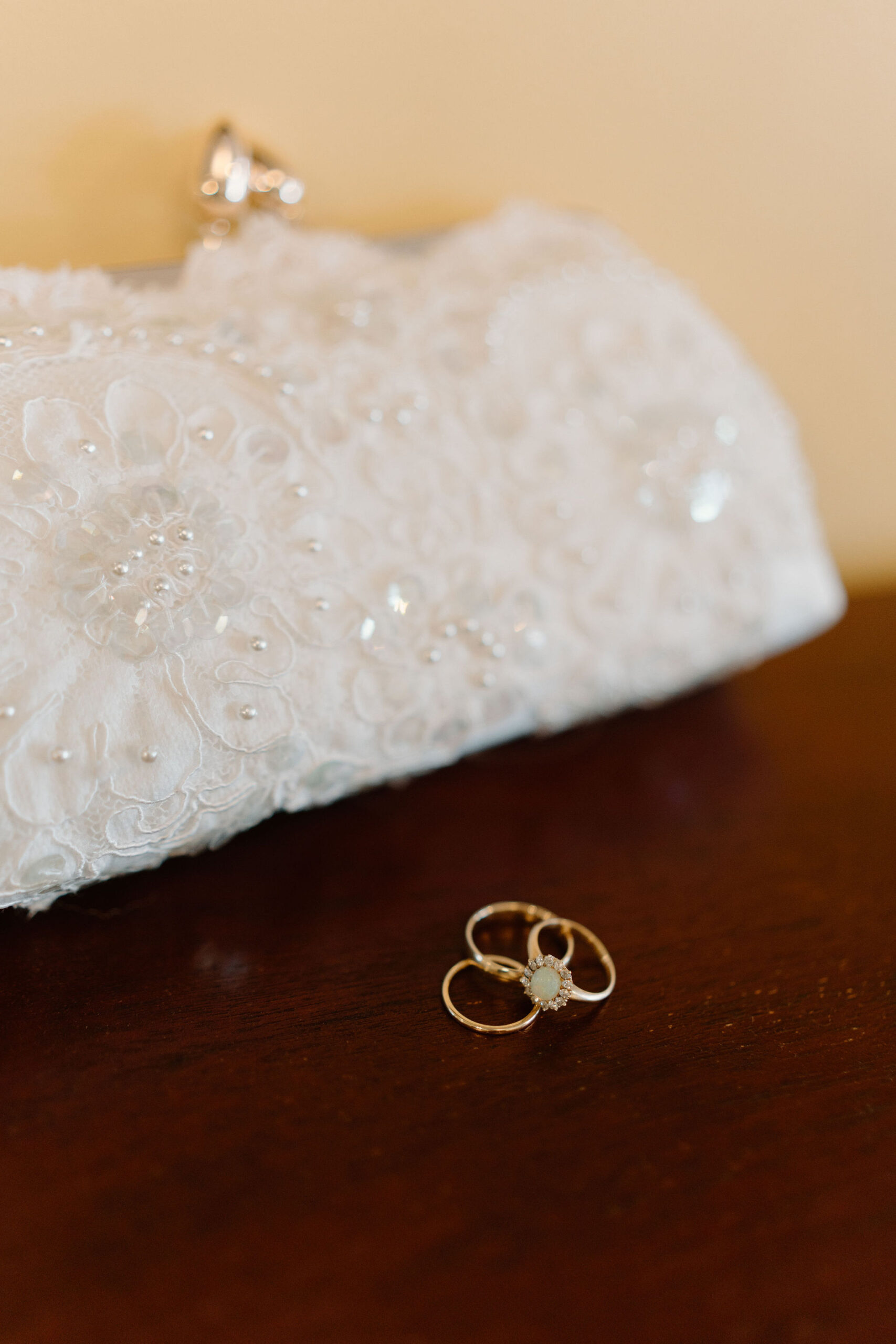 close up image of 2 gold wedding rings and 1 gold engagement ring with a pale green stone, displayed on a dark red wood, with a beaded white clutch in the background