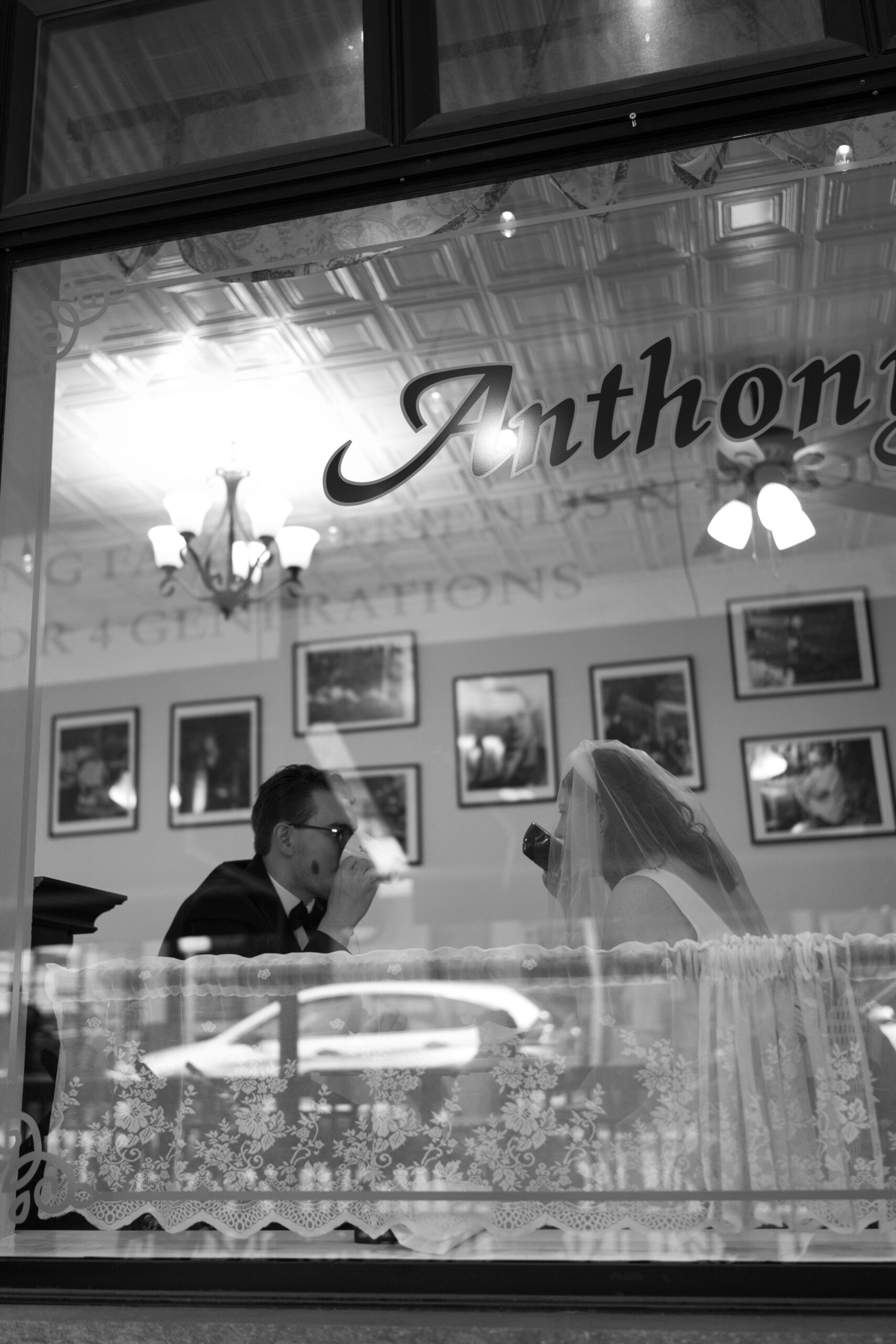 black and white image from exterior, looking through a window, of bride and groom seated at a table, each sipping their coffee