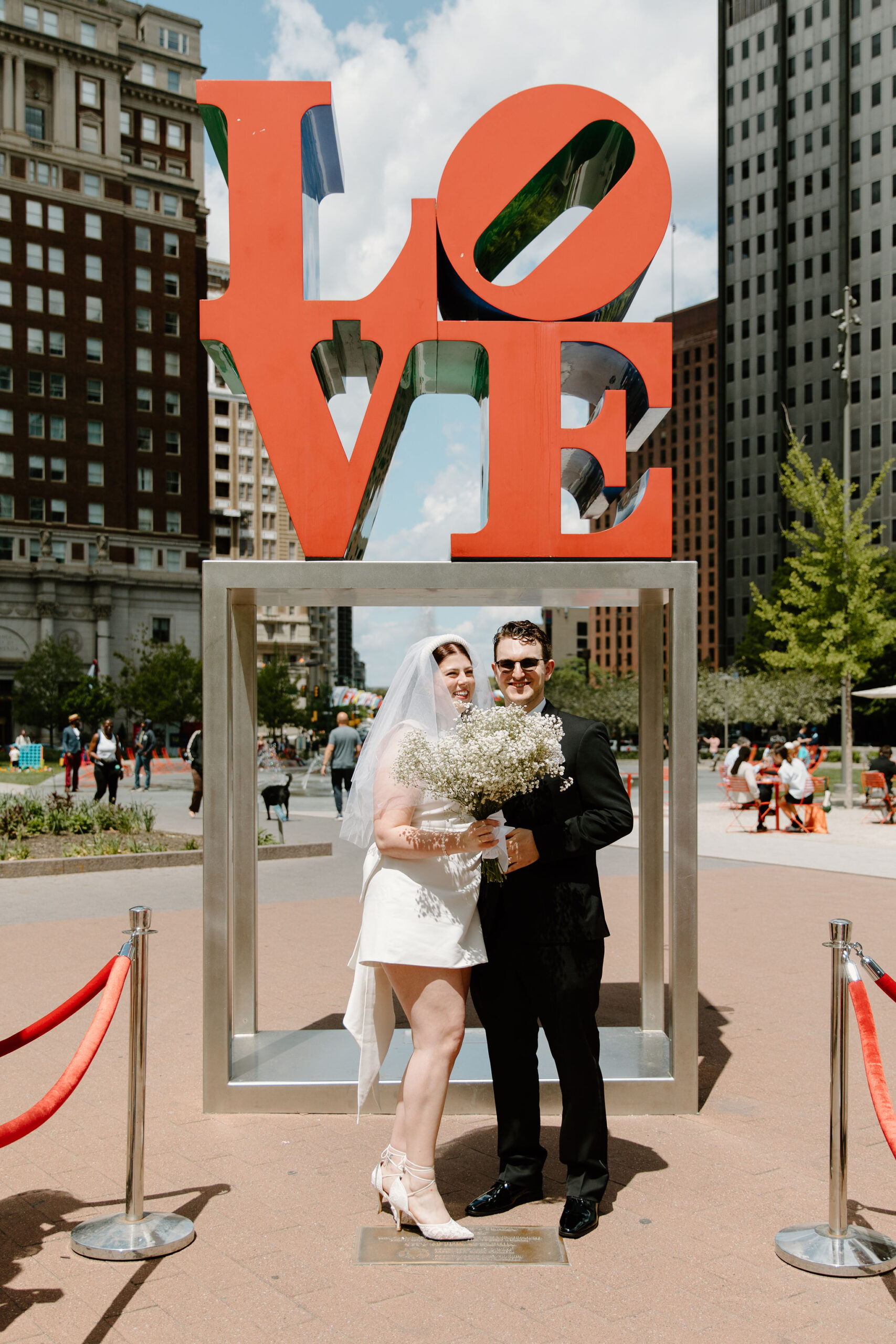 bride and groom smiling toward their guests, bride holding her bouquet, in front of the Love Park sign